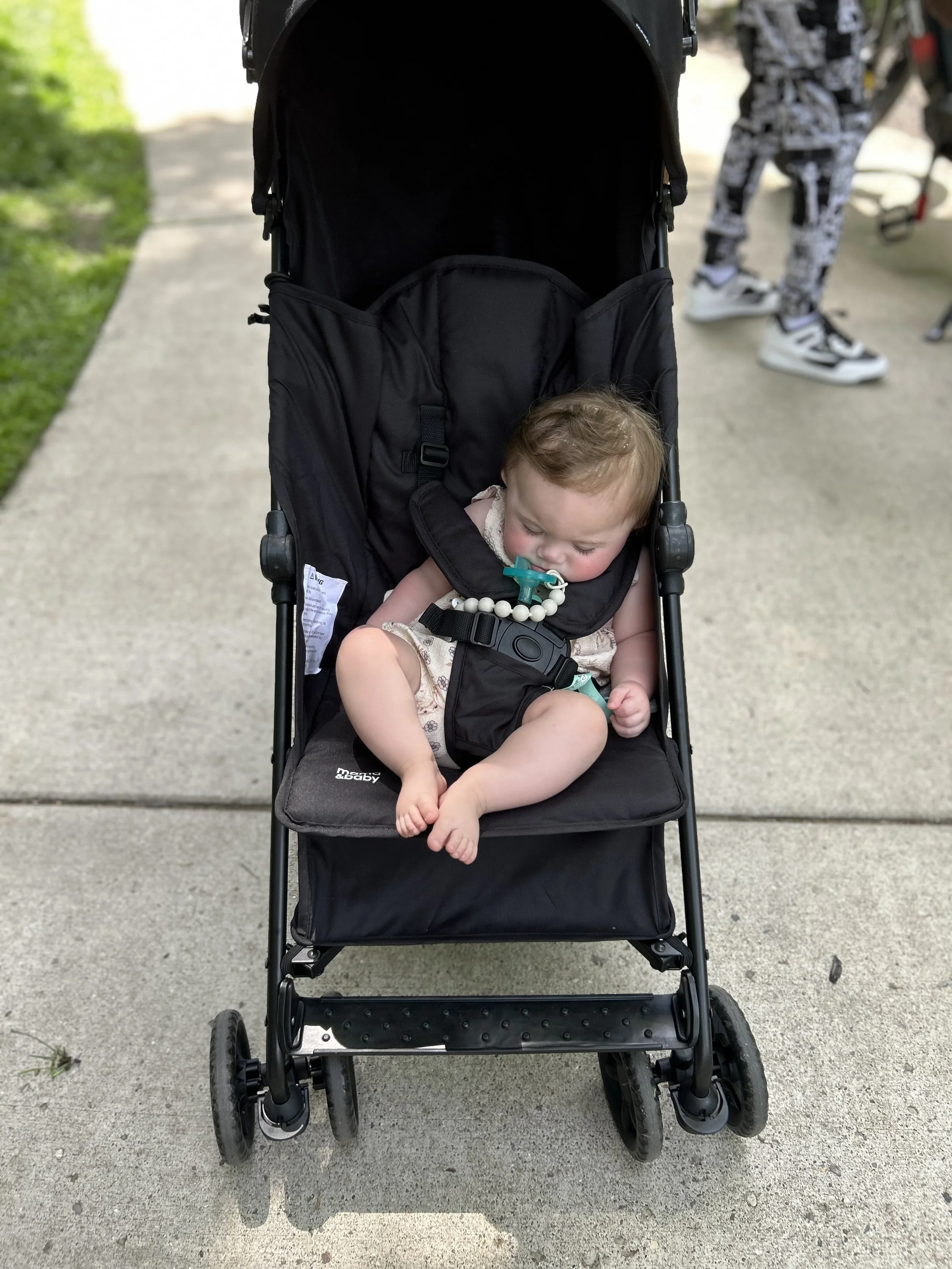 A baby sleeping in a black stroller on a sidewalk, holding a pacifier in their mouth and wearing a beaded necklace.