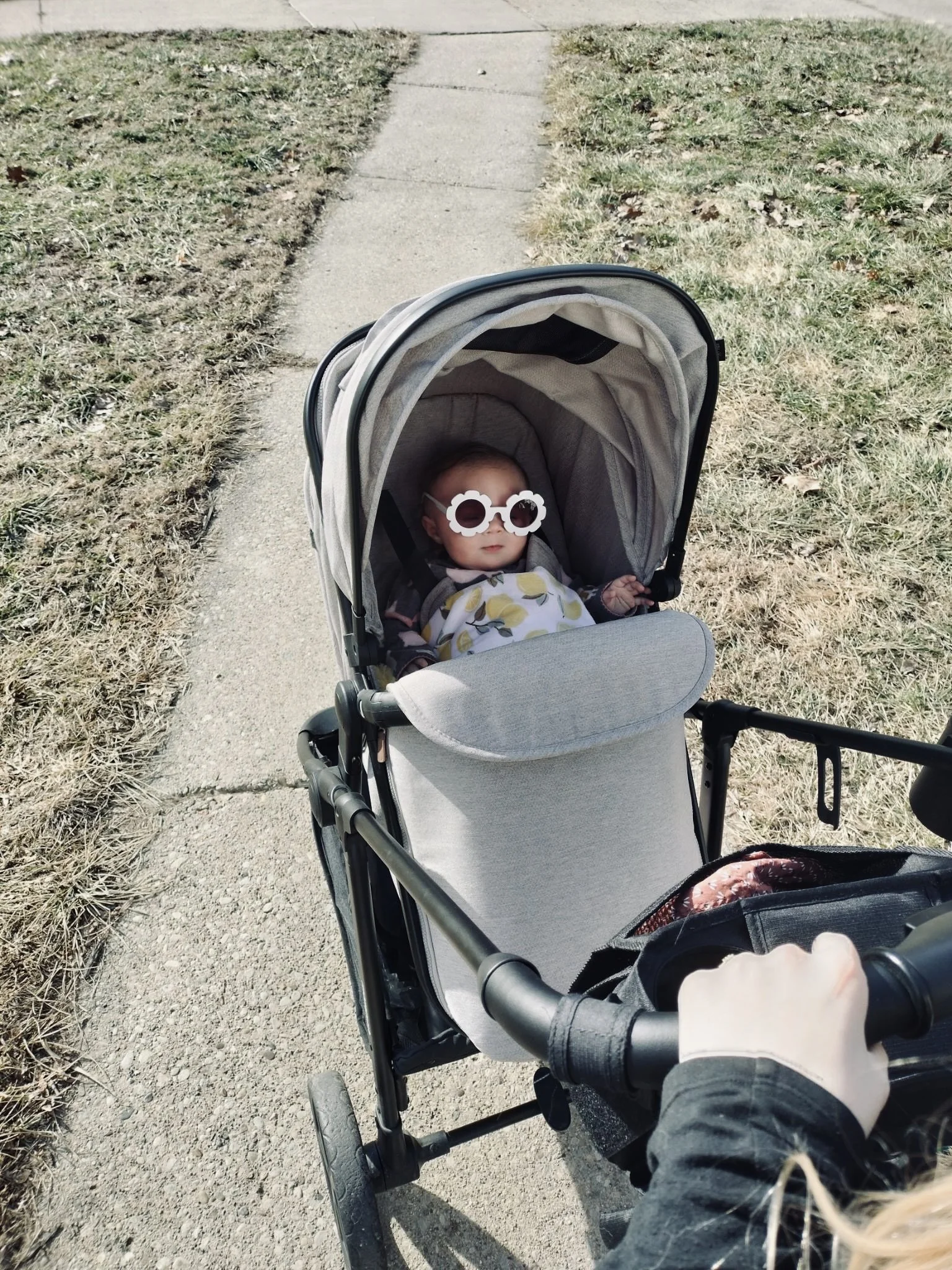 A baby wearing flower-shaped sunglasses sitting in a stroller on a sidewalk.