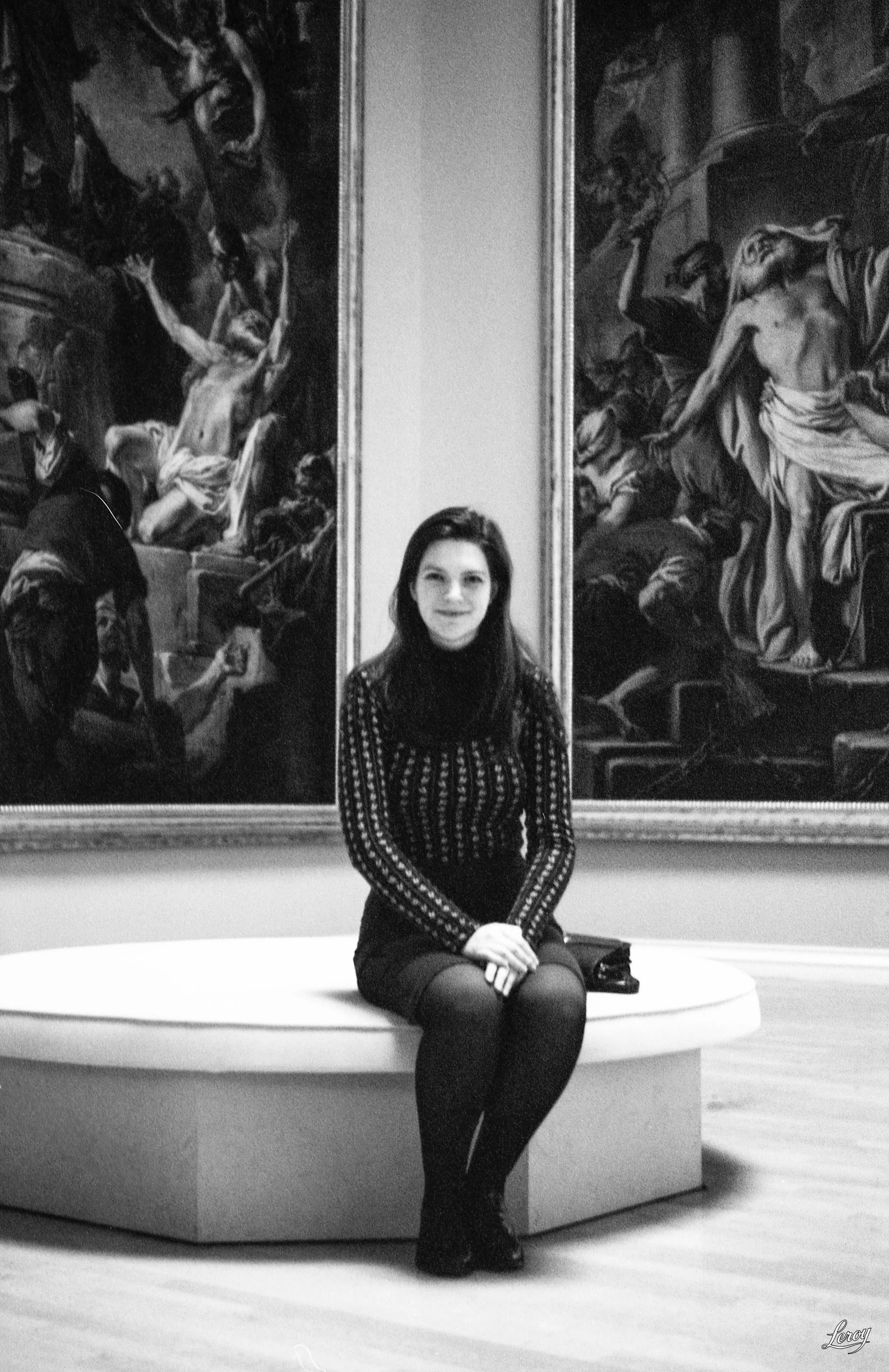 Une femme assise sur un banc dans un musée d'art, en noir et blanc, avec deux grands tableaux classiques en arrière-plan.