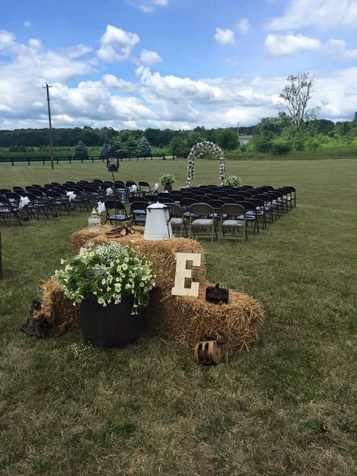 Outdoor wedding ceremony setup with rows of black chairs, a flower arch, hay bales decorated with flowers, letters, lanterns, and rustic decor on grassy field with cloudy sky.