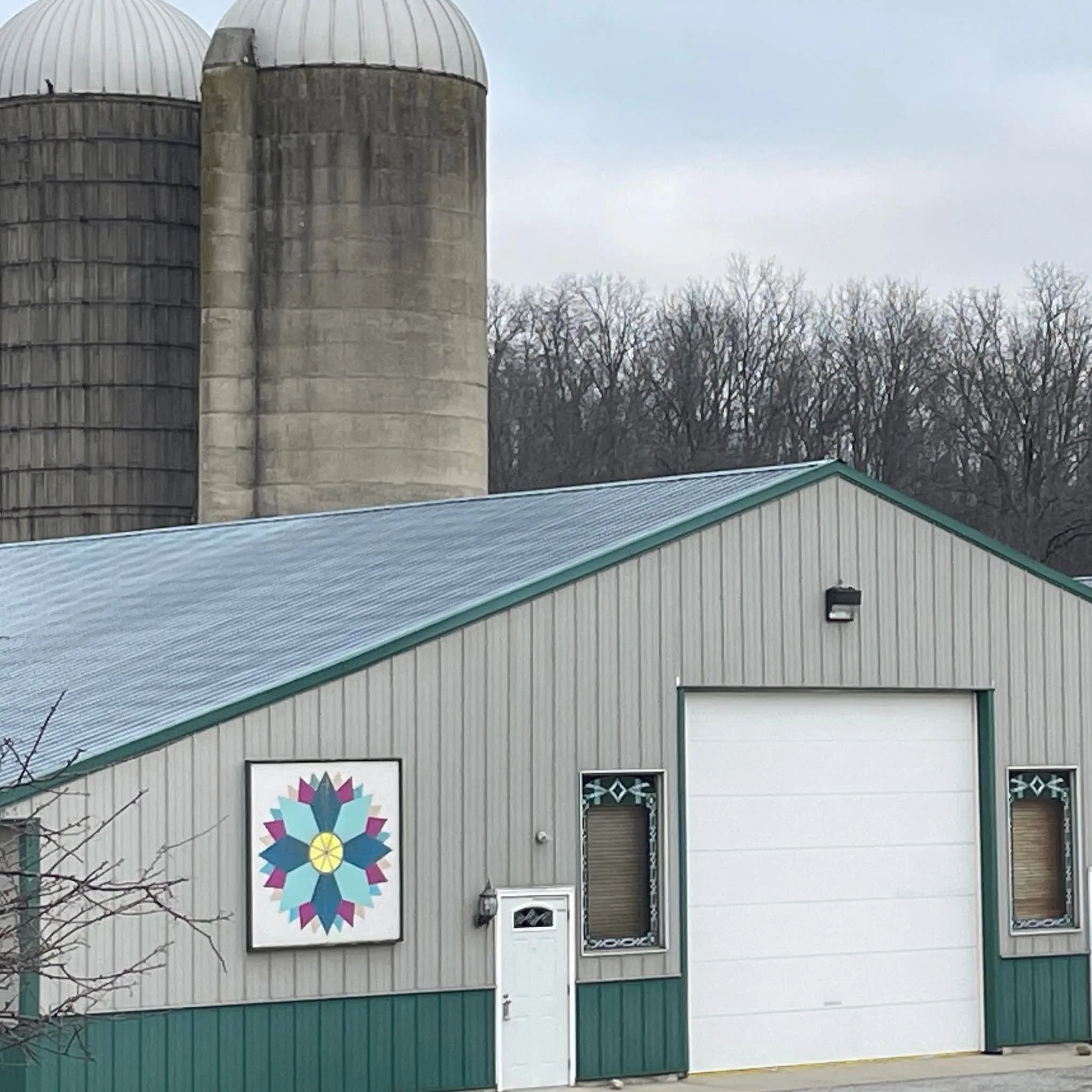 A grey metal building with a blue roof, decorated with a colorful geometric flower mural, and two small windows. In the background, there are three large concrete silos and leafless trees.