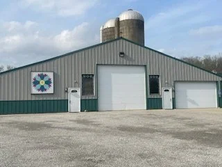 A large gray metal barn with two white garage doors and two trash bins in front. There is a colorful flower-shaped sign on the left side of the barn and silos on top.