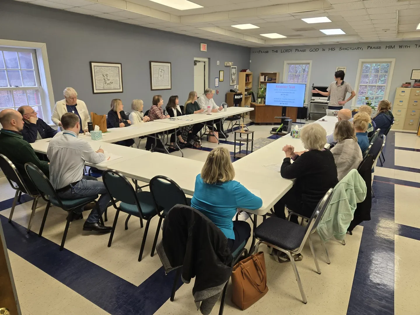 Group of people attending a presentation or meeting in a conference room with U-shaped tables, a presenter at the front, and a screen displaying a slide titled "Volunteer Team".