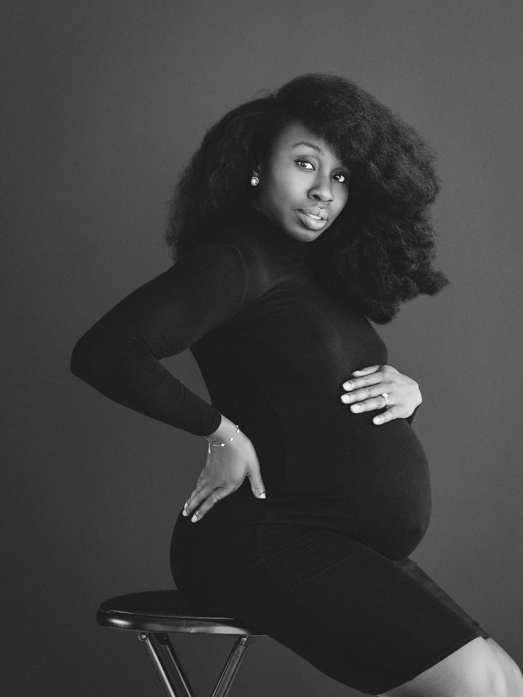 Black and white portrait of a pregnant woman sitting on a stool with one hand on her hip and the other on her belly, looking confidently at the camera.