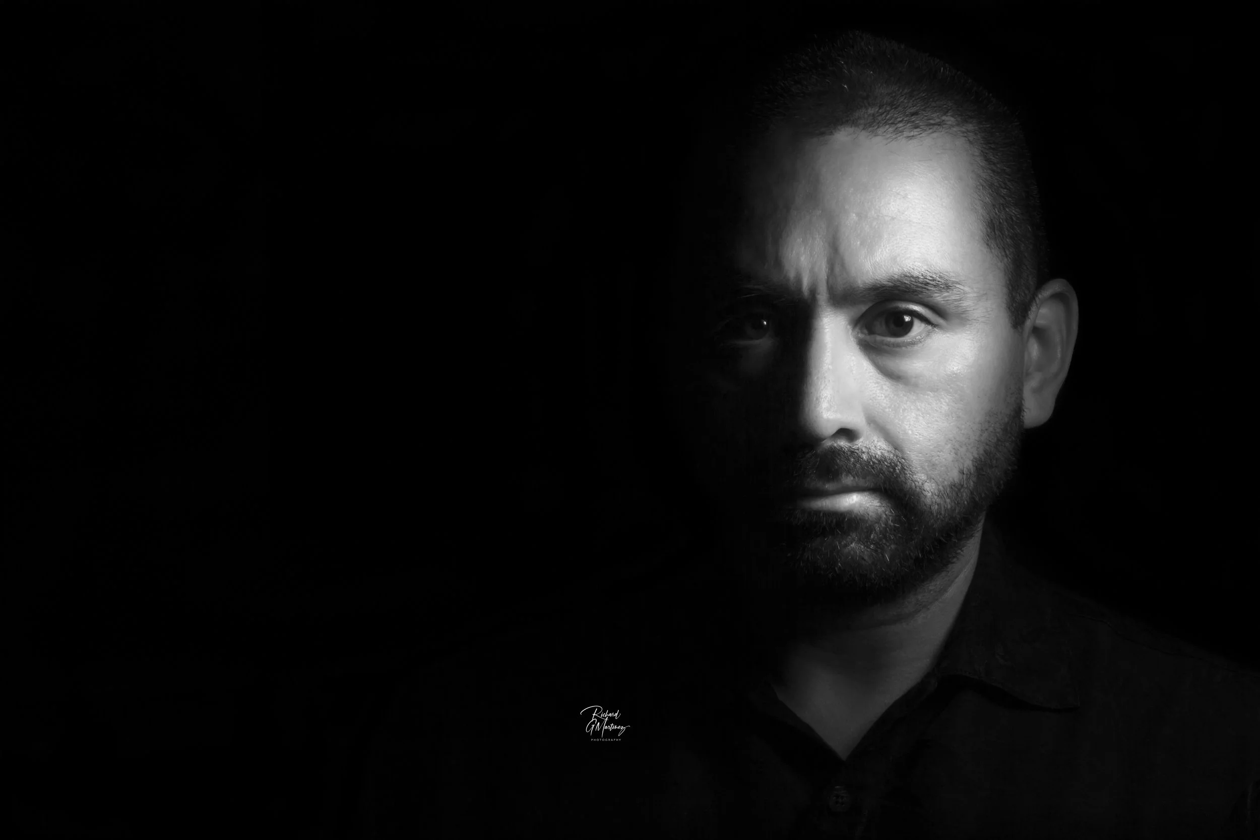 Black and white portrait of a man with short hair and a beard, lighting illuminating only the right side of his face, while the left side is in shadow.