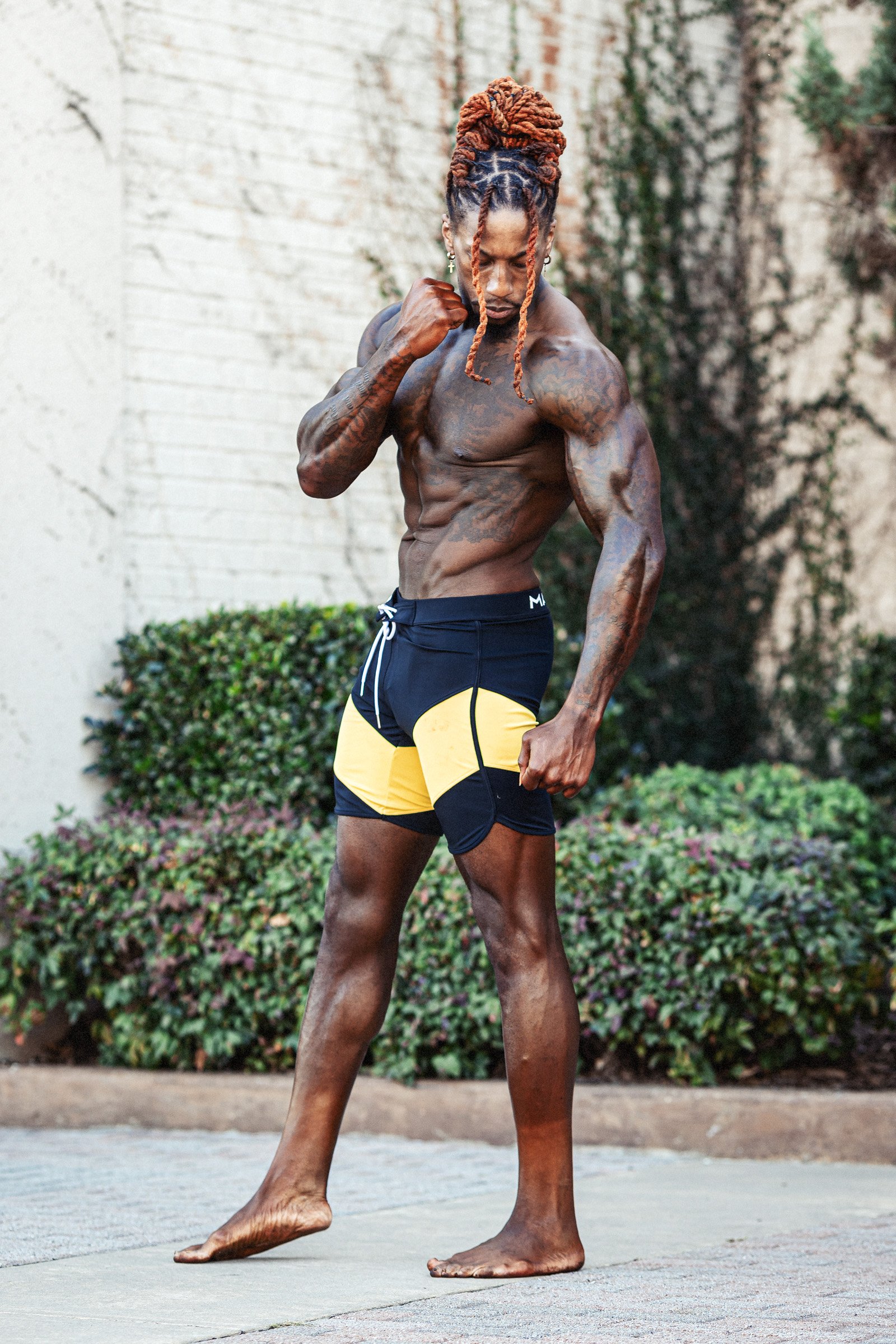A muscular man with tattoos and dreadlocks poses outdoors, flexing his arm, wearing black and yellow shorts.