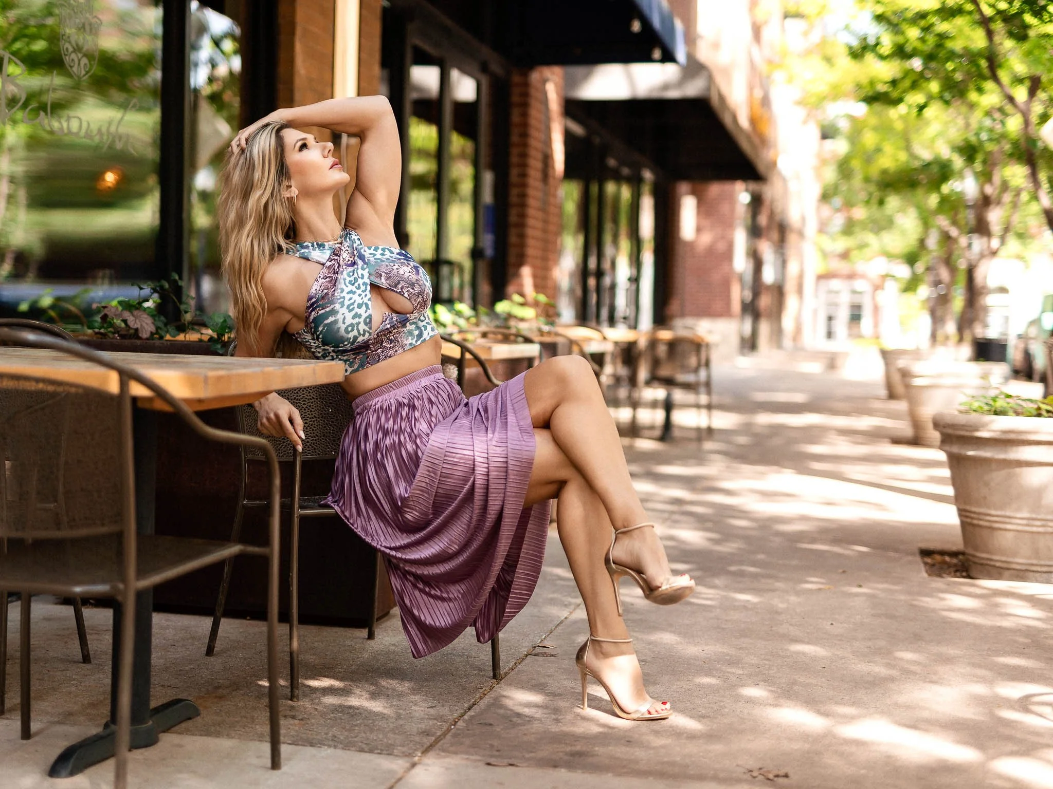 A woman with long wavy blonde hair, sitting on a chair outside a cafe, wearing a colorful sleeveless top, a purple pleated skirt, and beige high-heeled sandals, with one arm raised and her hand resting on her head, looking relaxed and stylish.