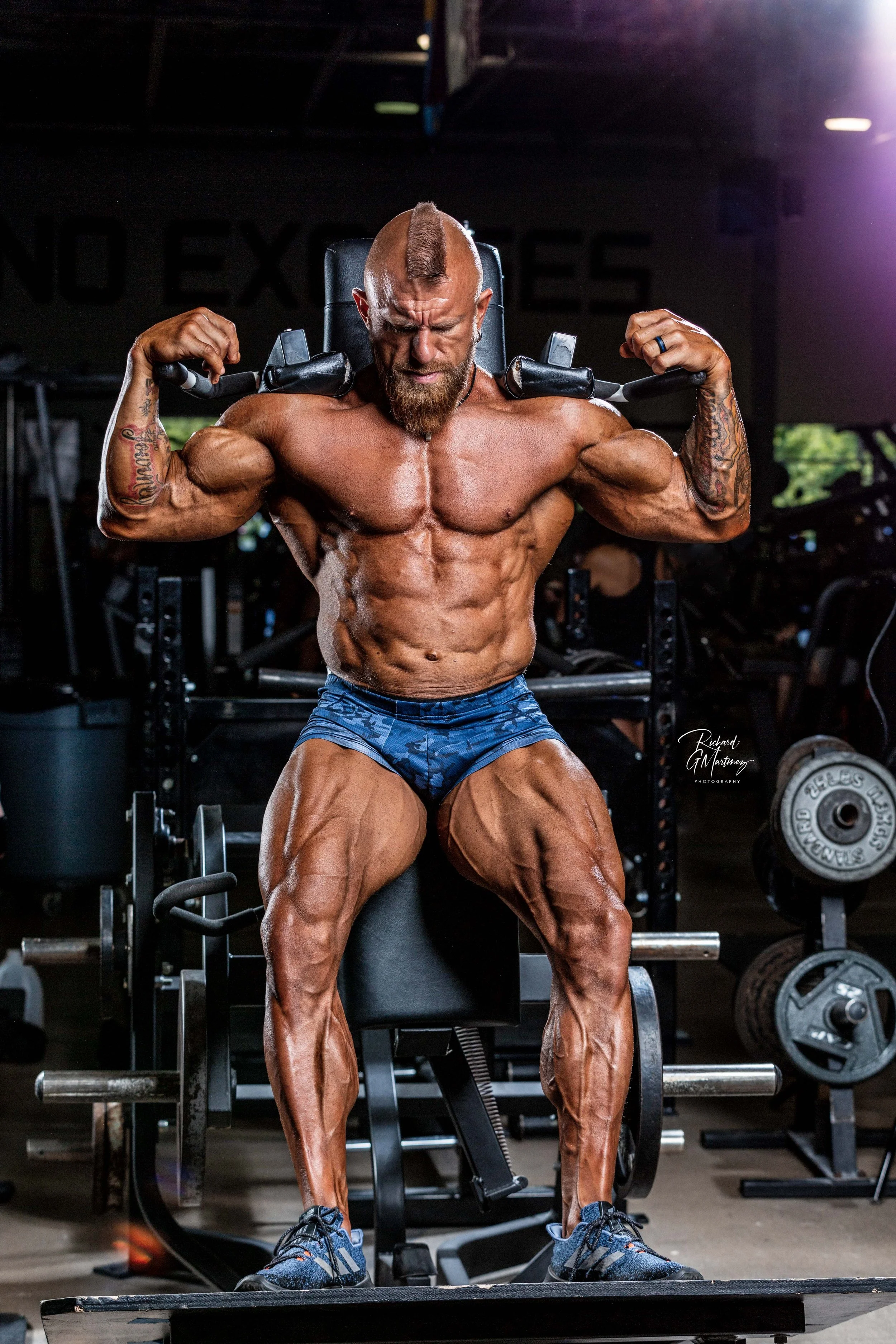 A muscular man with a mohawk hairstyle and tattoos on his arms, wearing blue shorts and athletic shoes, is sitting on a workout bench in a gym, flexing his biceps.