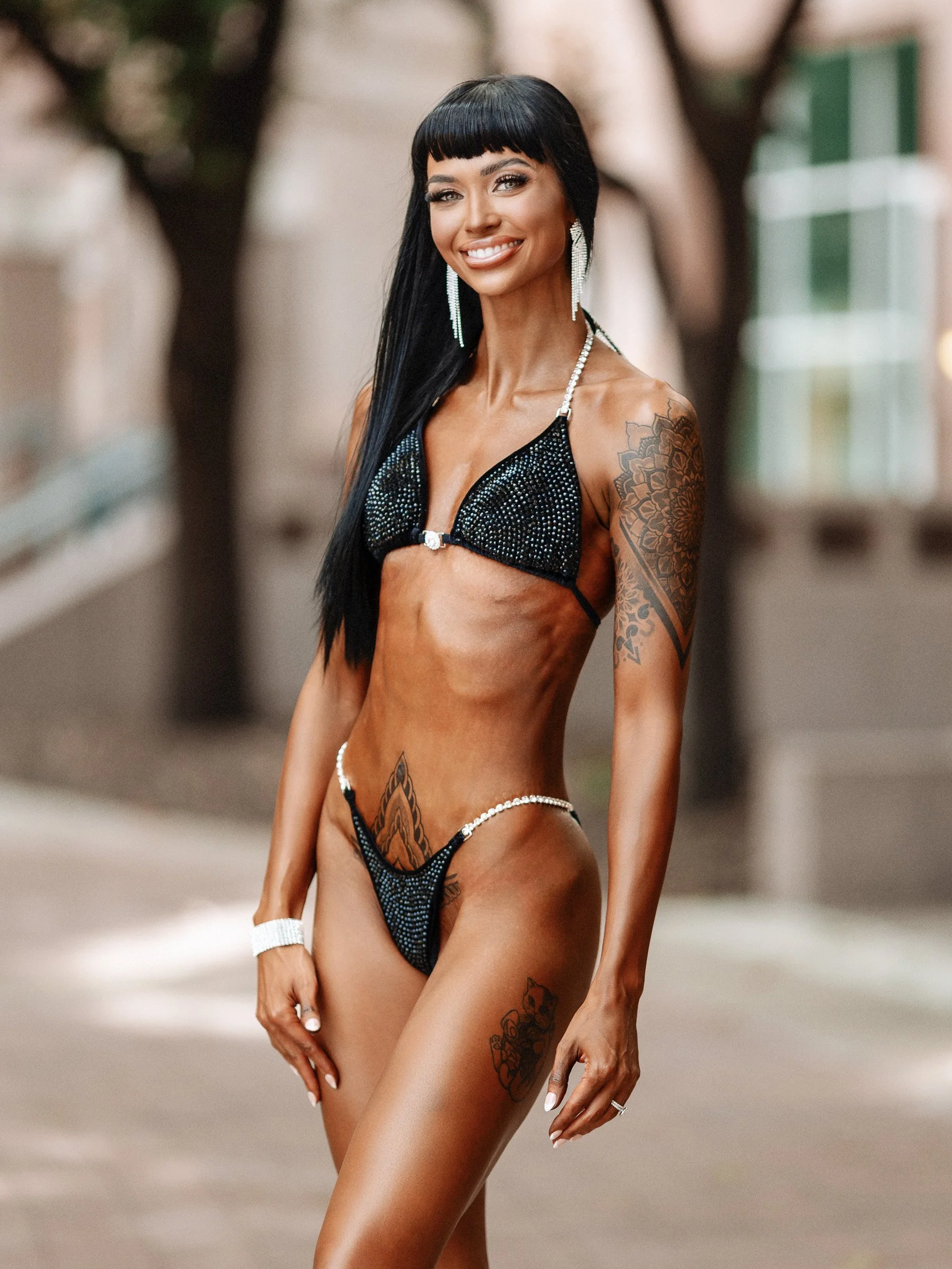 A woman with long black hair, tattoos, and a fit physique wearing a rhinestone-studded dark bikini, standing outdoors with trees and buildings in the background, smiling at the camera.
