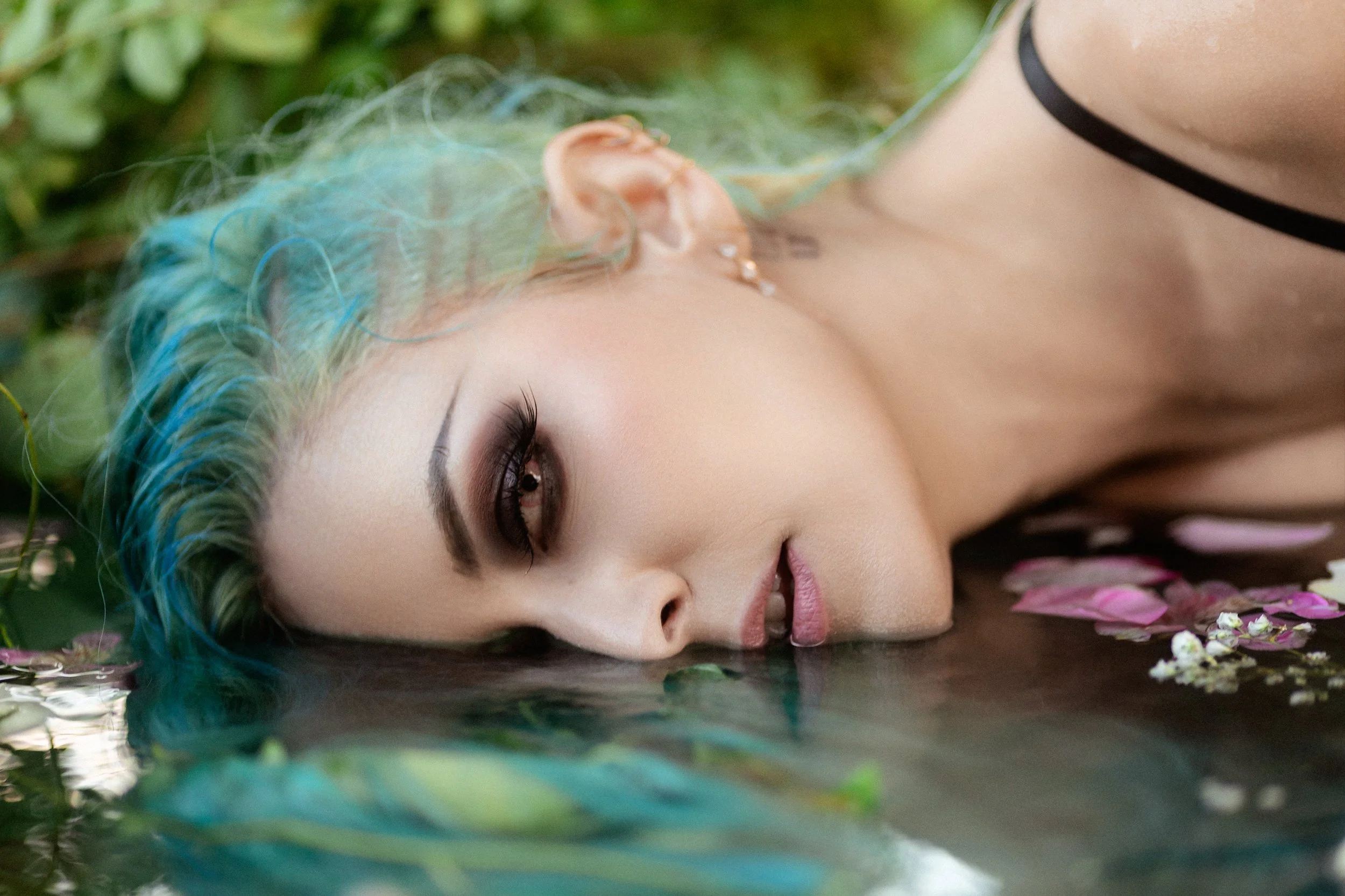 A young woman with vibrant teal and green hair, lying on her side by the water, with her face close to the surface and her eyes closed, surrounded by pink and white flowers floating on the water.