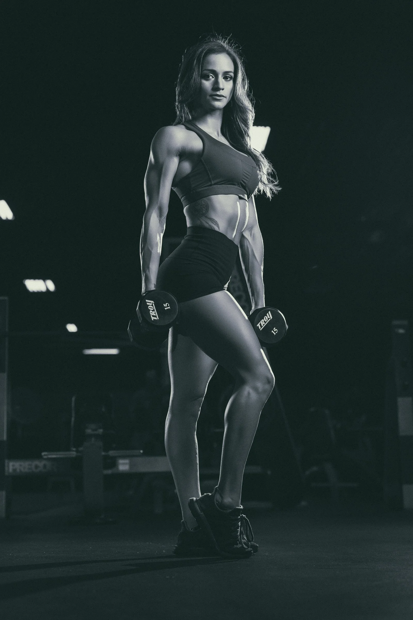 IFBB bikini pro posing with dumb bells looking toward the camera in a black and white image