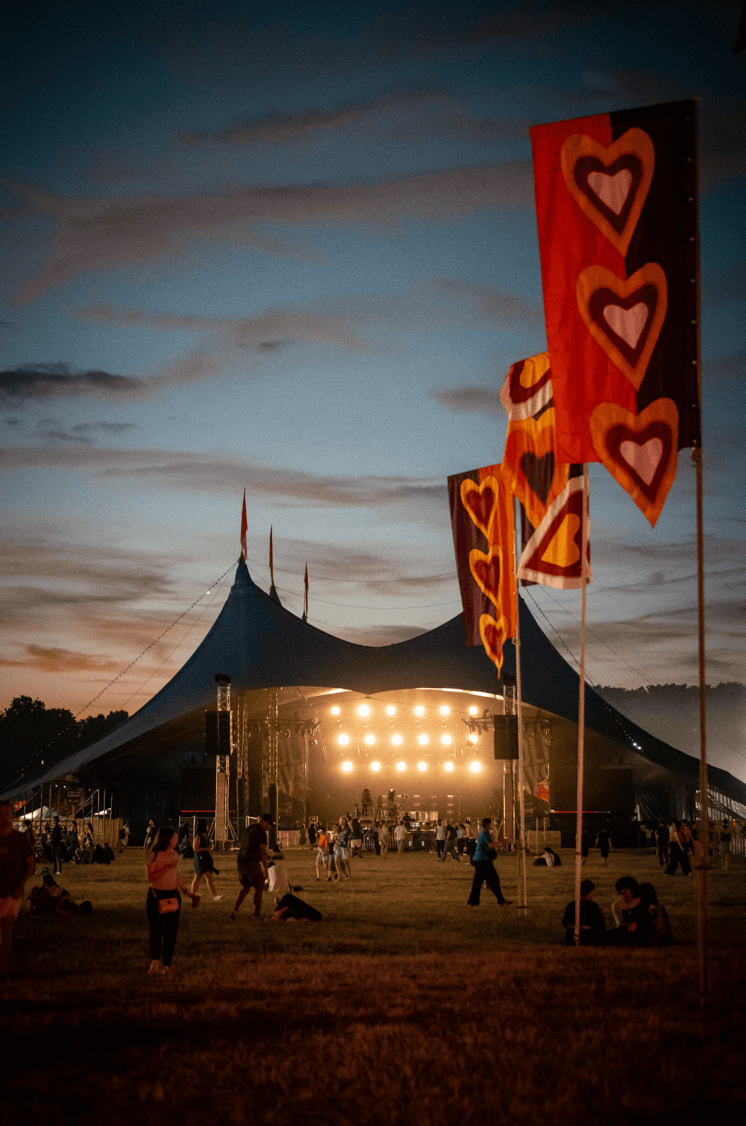 Un grand chapiteau de festival en soirée avec des drapeaux colorés en forme de cœurs devant, des lumières sur scène, et des gens assis ou marchant sur l'herbe.