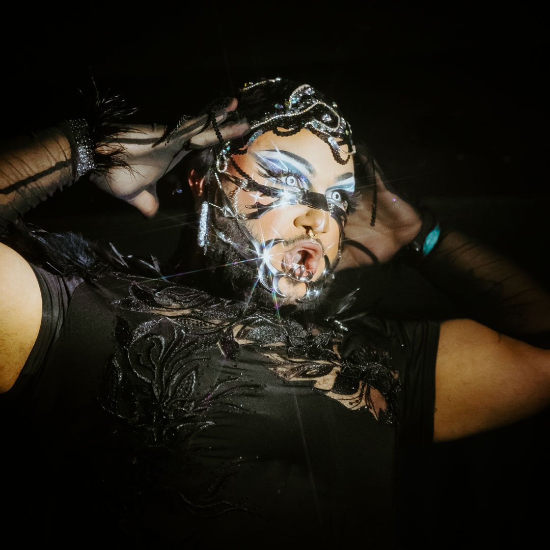 Une personne avec un maquillage artistique, portant des accessoires brillants et un costume noir, repose dans un environnement sombre.