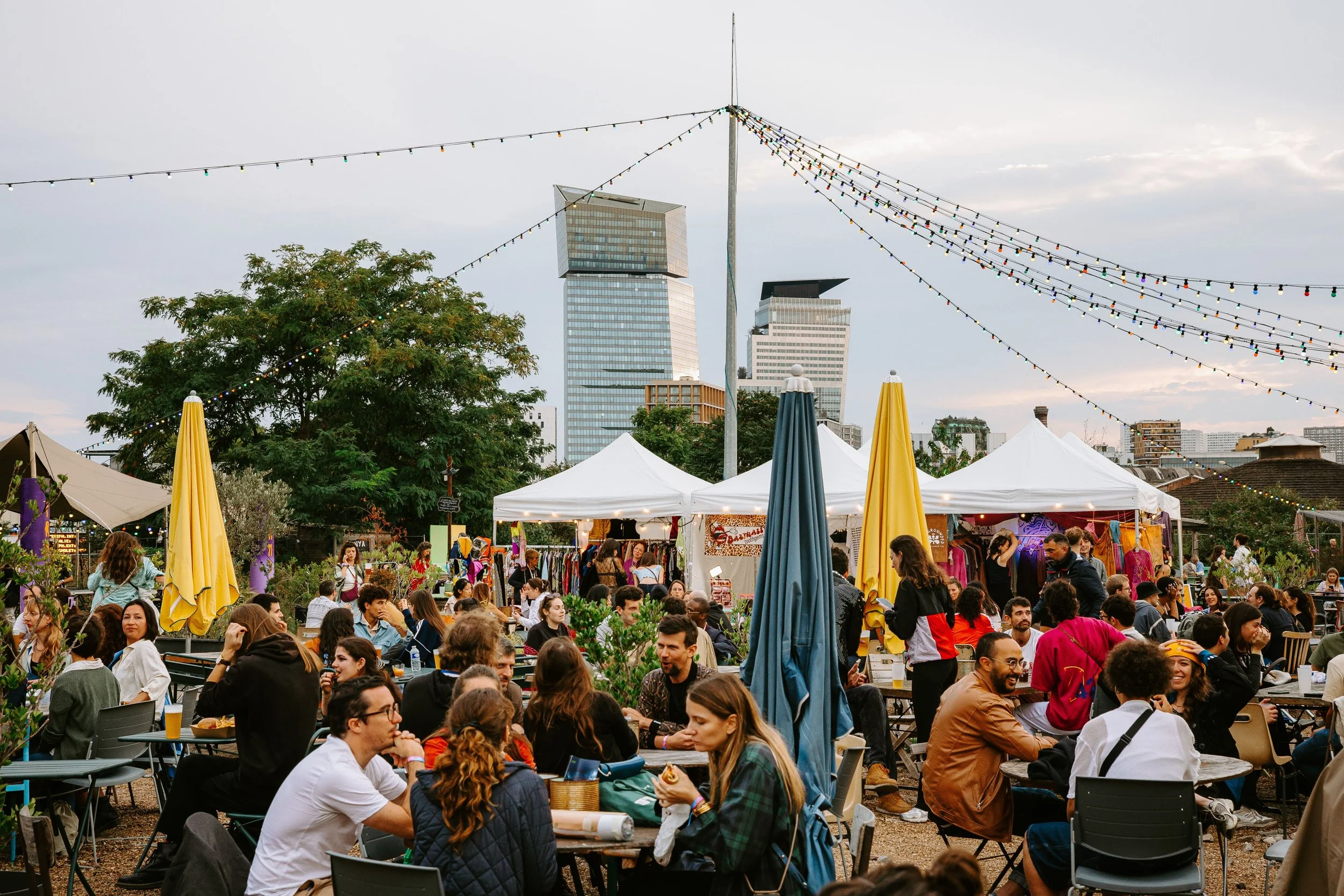 Plan large du Festival Fluctuations. On y voit une foule de gens assises et debout en plein air avec des stands, des parasols jaunes et bleus, des décorations lumineuses suspendues, un gratte-ciel en arrière-plan.