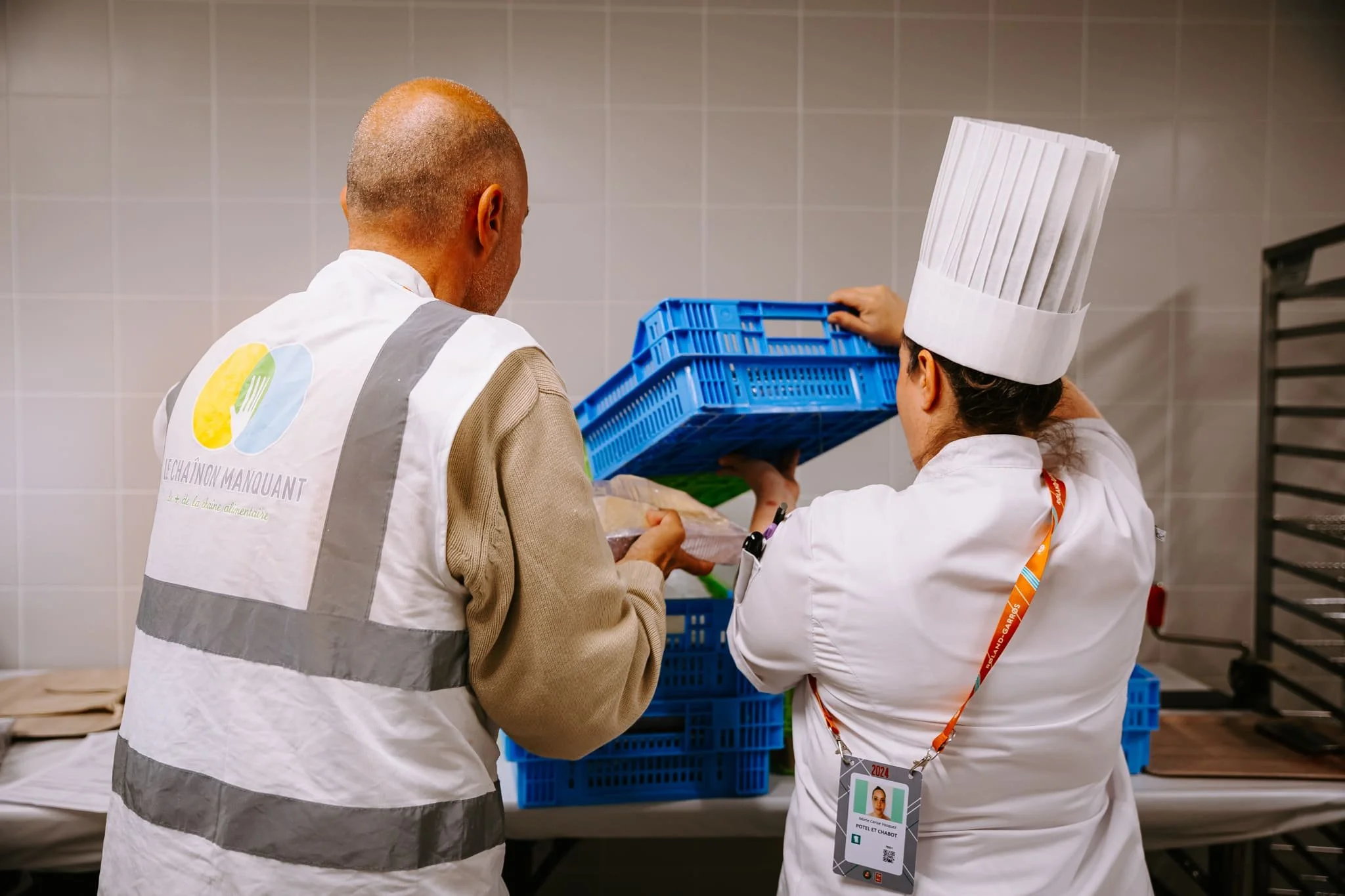 Photo d'une cheffe cuisinière en uniforme donnant une caisse de nourriture non-consommée à un bénévole du Chaînon Manquant, qui porte un veston floqué du logo de l'association.