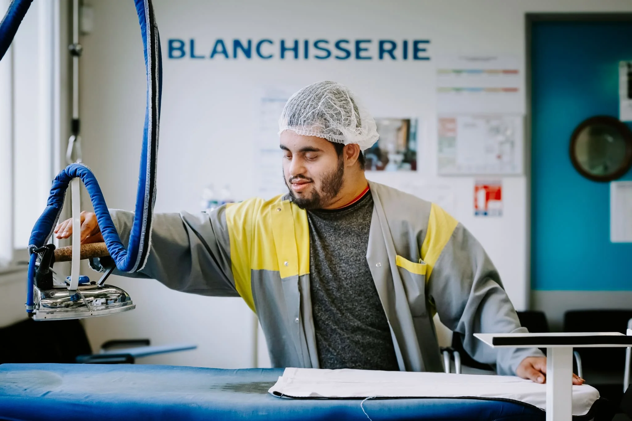 Photo prise au sein de l'ESAT le Castel, un homme en situation de handicap portant une blouse de travail et un bonnet de protection, en train de repasser un vêtement dans un atelier de blanchisserie.