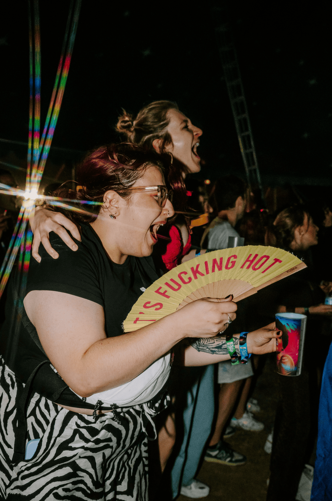Deux femmes rient et s'amusent lors d'un festival ou concert, une tient un éventail avec l'inscription 'Fucking Hot'.