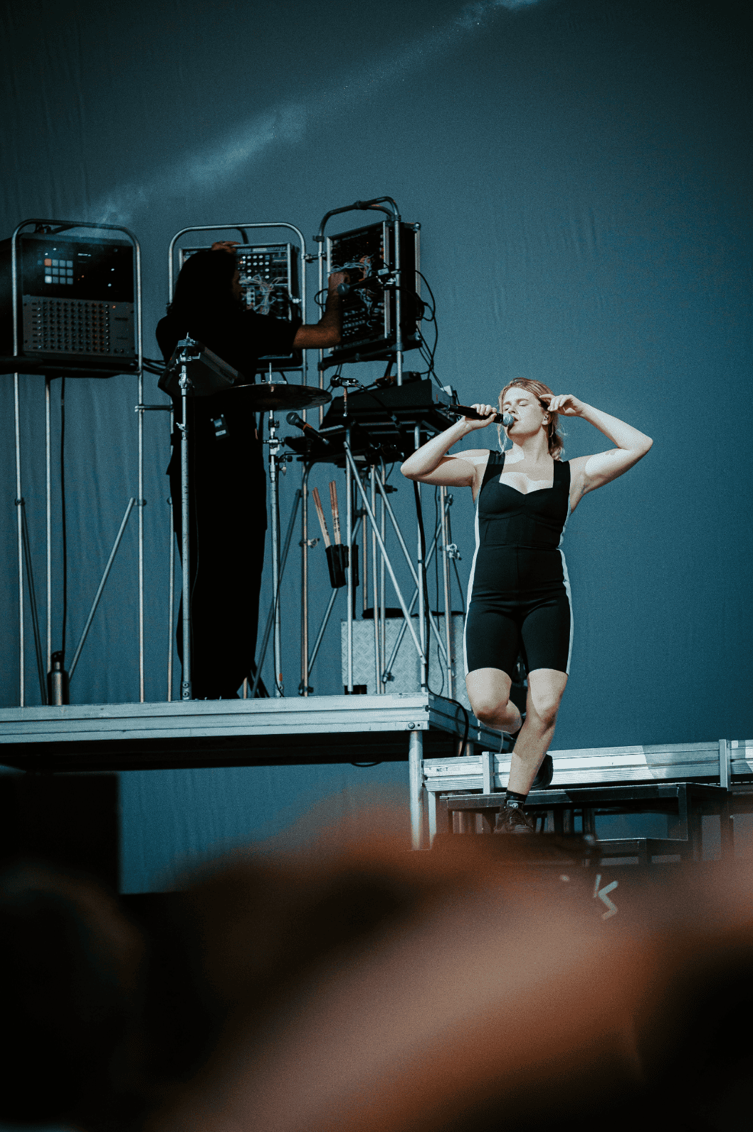 Une femme chante avec un microphone sur scène, tandis qu'une autre personne manipule du matériel électronique en arrière-plan.