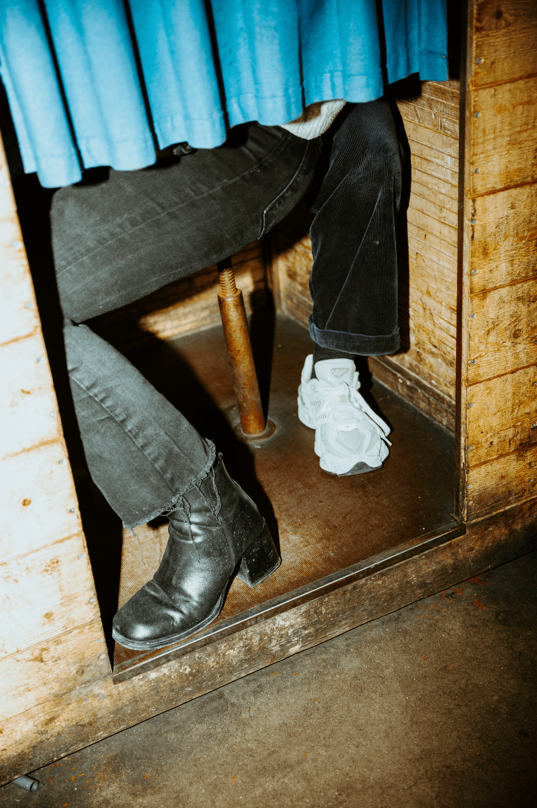 Photo de deux personnes assises l'une derrière l'autre dans un photomaton. On ne devine que leurs jambes et leurs pieds : une personne porte des bottines noires à talons, l'autre des baskets blanches.