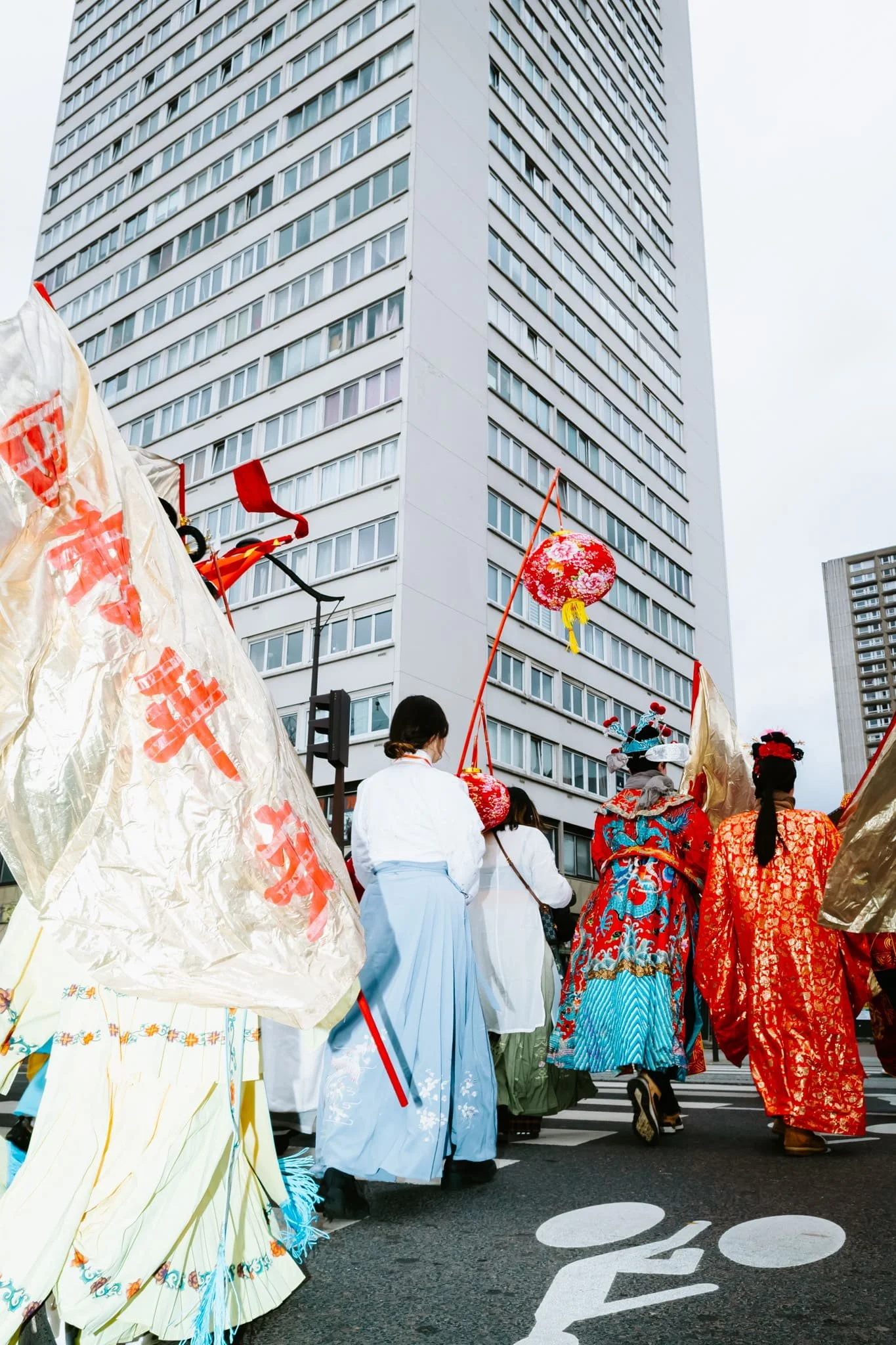 Photo d'un groupe de personnes en costumes traditionnels avançant dans la rue lors de la parade du Nouvel an chinois à Paris 13e, avec un grand immeuble moderne en arrière-plan.