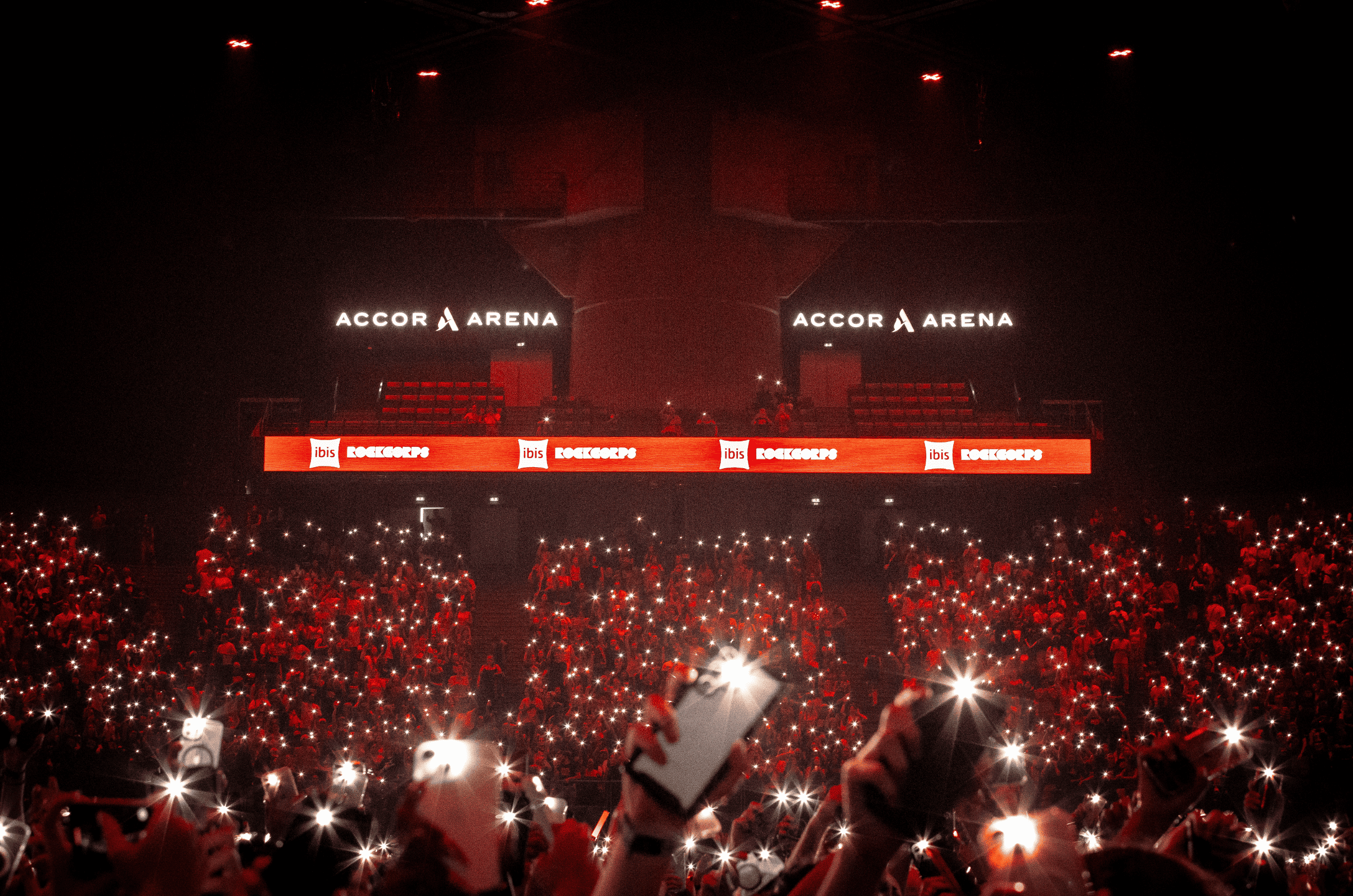 Concert à l'intérieur de l'Aréna d'Accor avec des spectateurs utilisant leurs téléphones portables pour faire des lumières, écrans affichant 'ibis ROCKCORPS', et un grand écran lumineux.