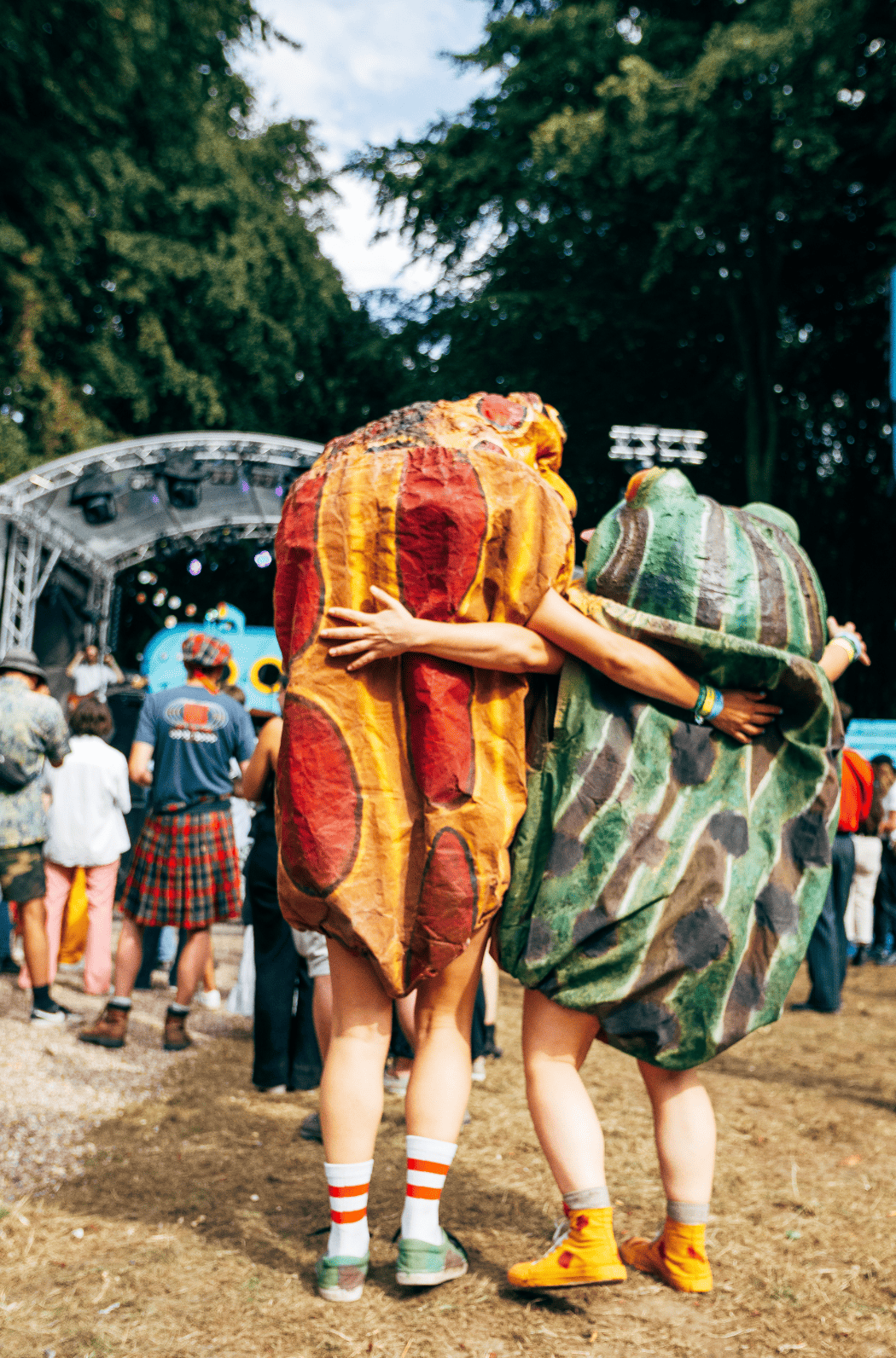 Deux personnes déguisées en légumes, une en poivron rouge et l'autre en courgette, se donnent une accolade lors d'un festival en plein air, avec une scène et un public en arrière-plan.