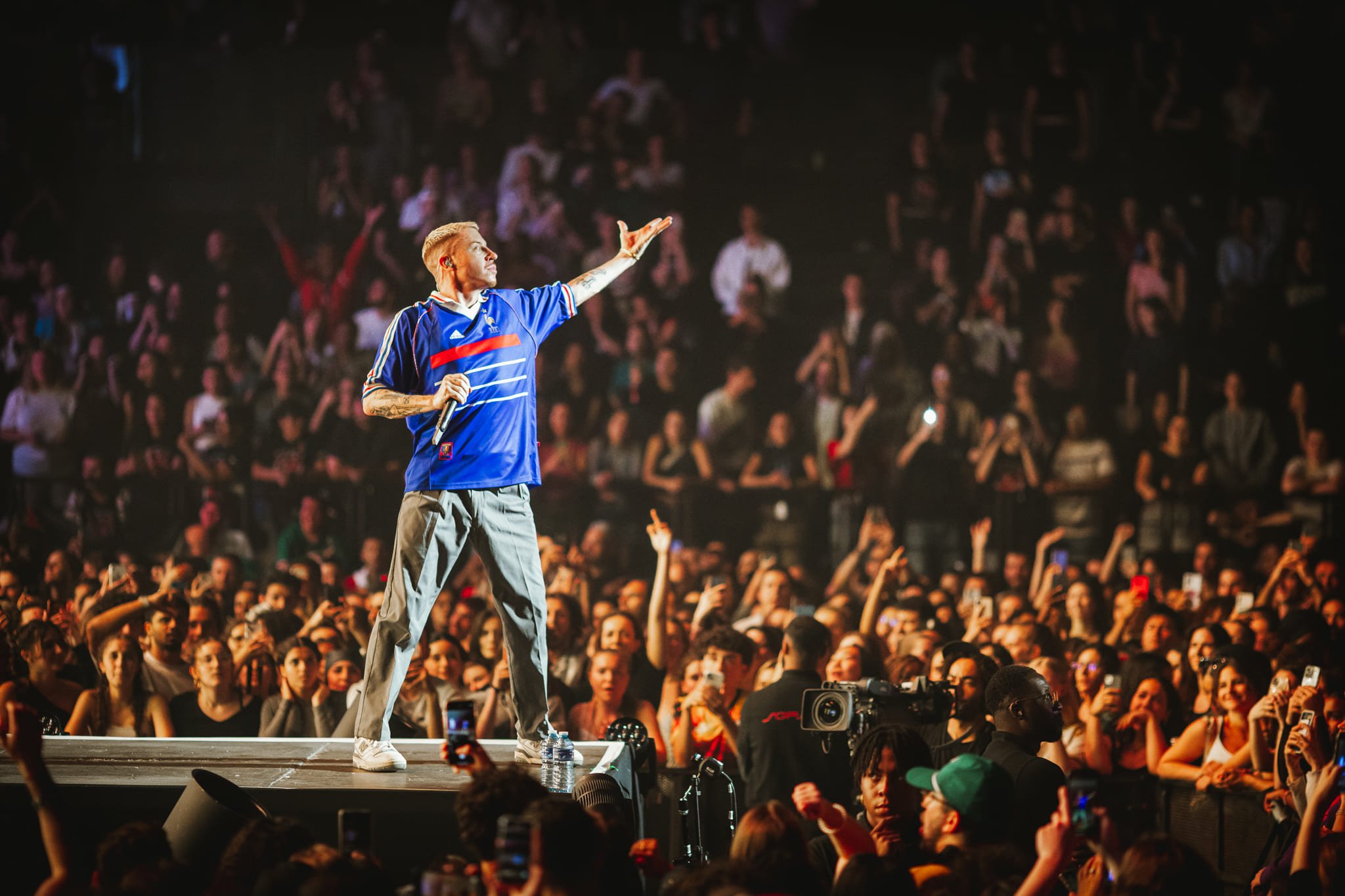 Un artiste debout sur scène chante devant une grande foule lors d'un concert, utilisant un micro, avec des fans prenant des photos ou vidéos.