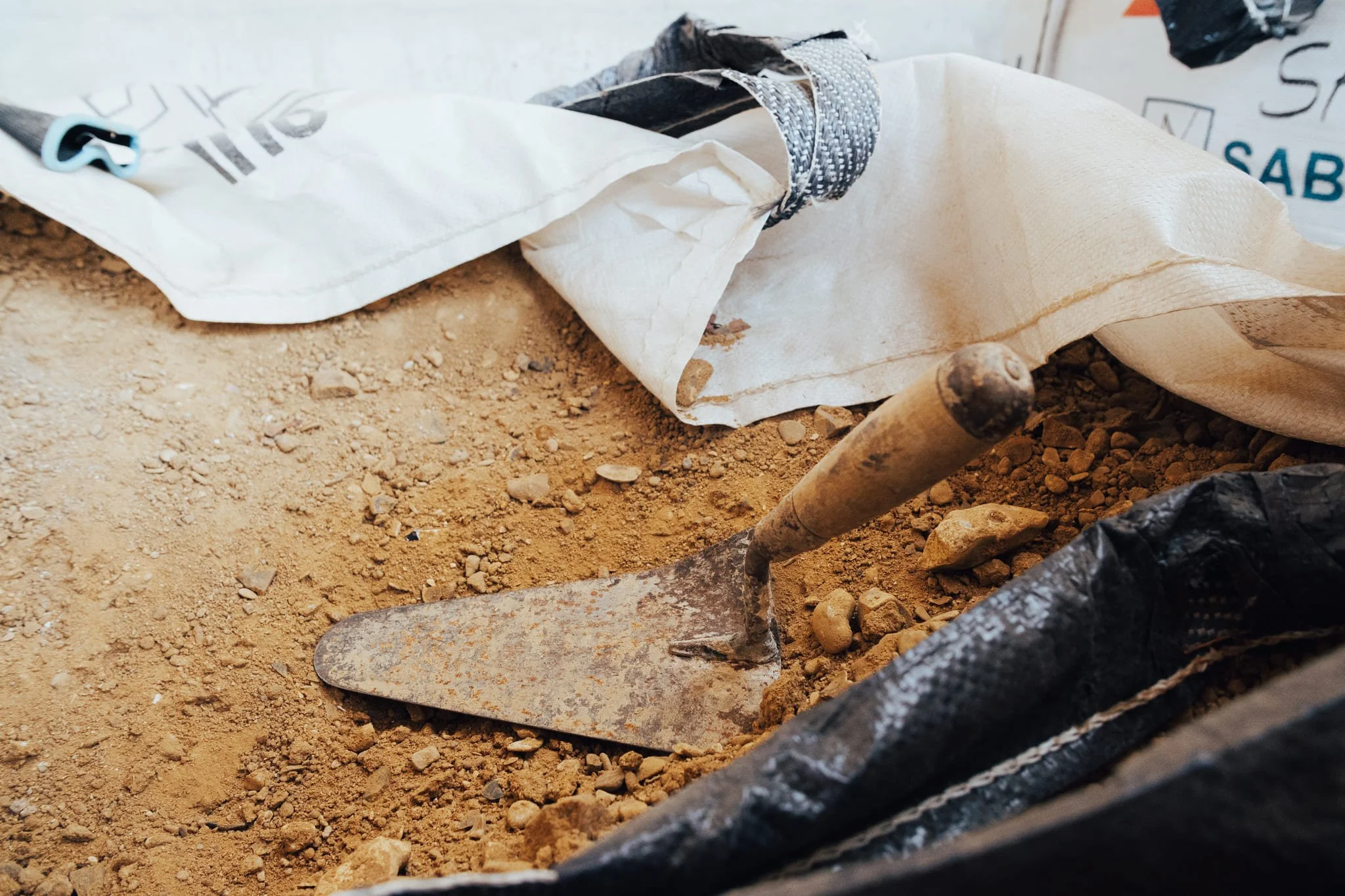 Une pelle de chantier enfoncée dans le sol de terre sèche entourée de sacs de matériaux de construction.
