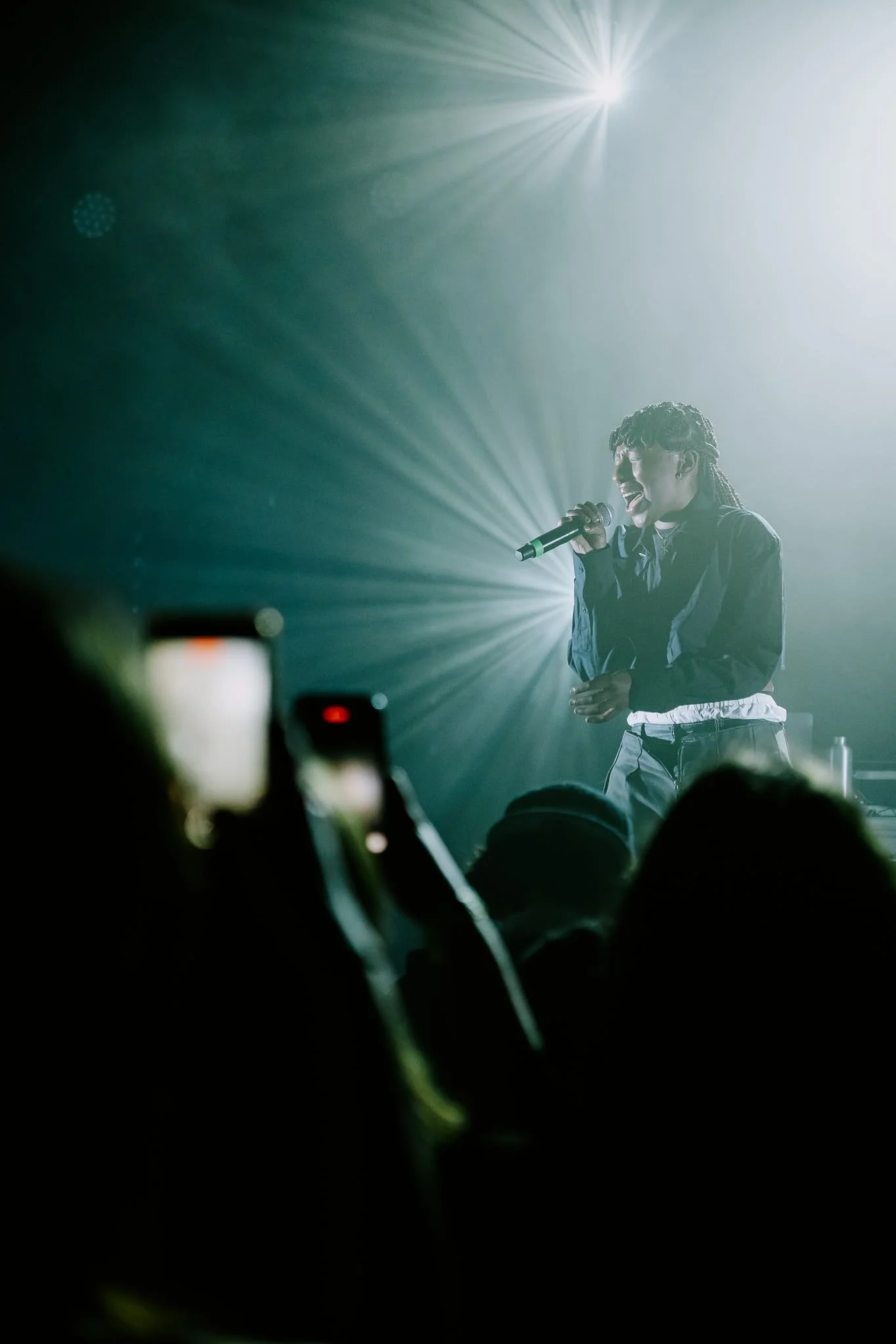Une chanteuse sur scène avec un micro, éclairée par des projecteurs lumineux, entourée de spectateurs, certains prenant des photos avec leur téléphone.