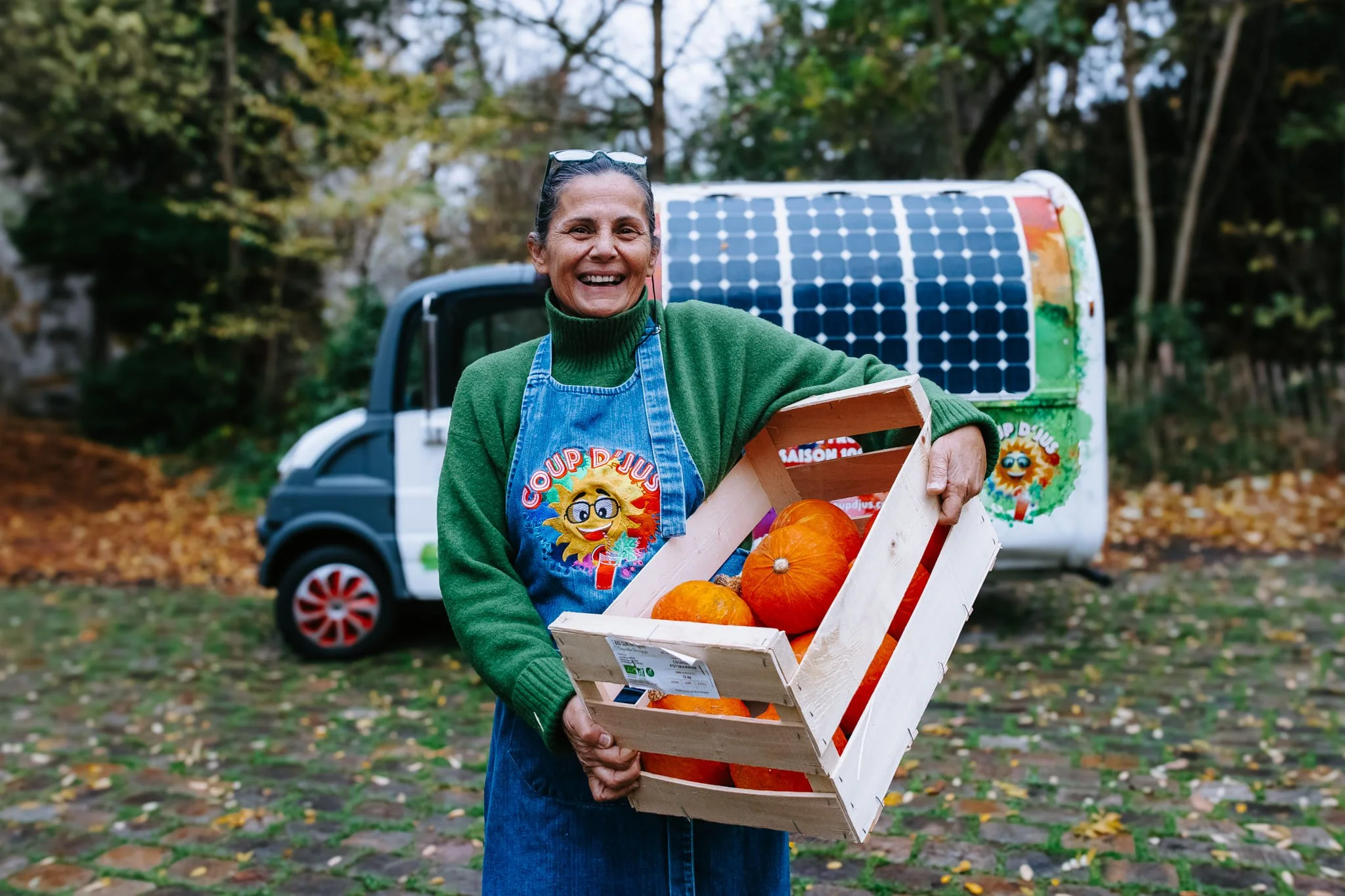 Reportage photo auprès de Coup'd'Jus, un foodtruck engagé. Une femme souriante tient une caisse en bois remplie de courges devant son foodtruck équipé de panneaux solaires.