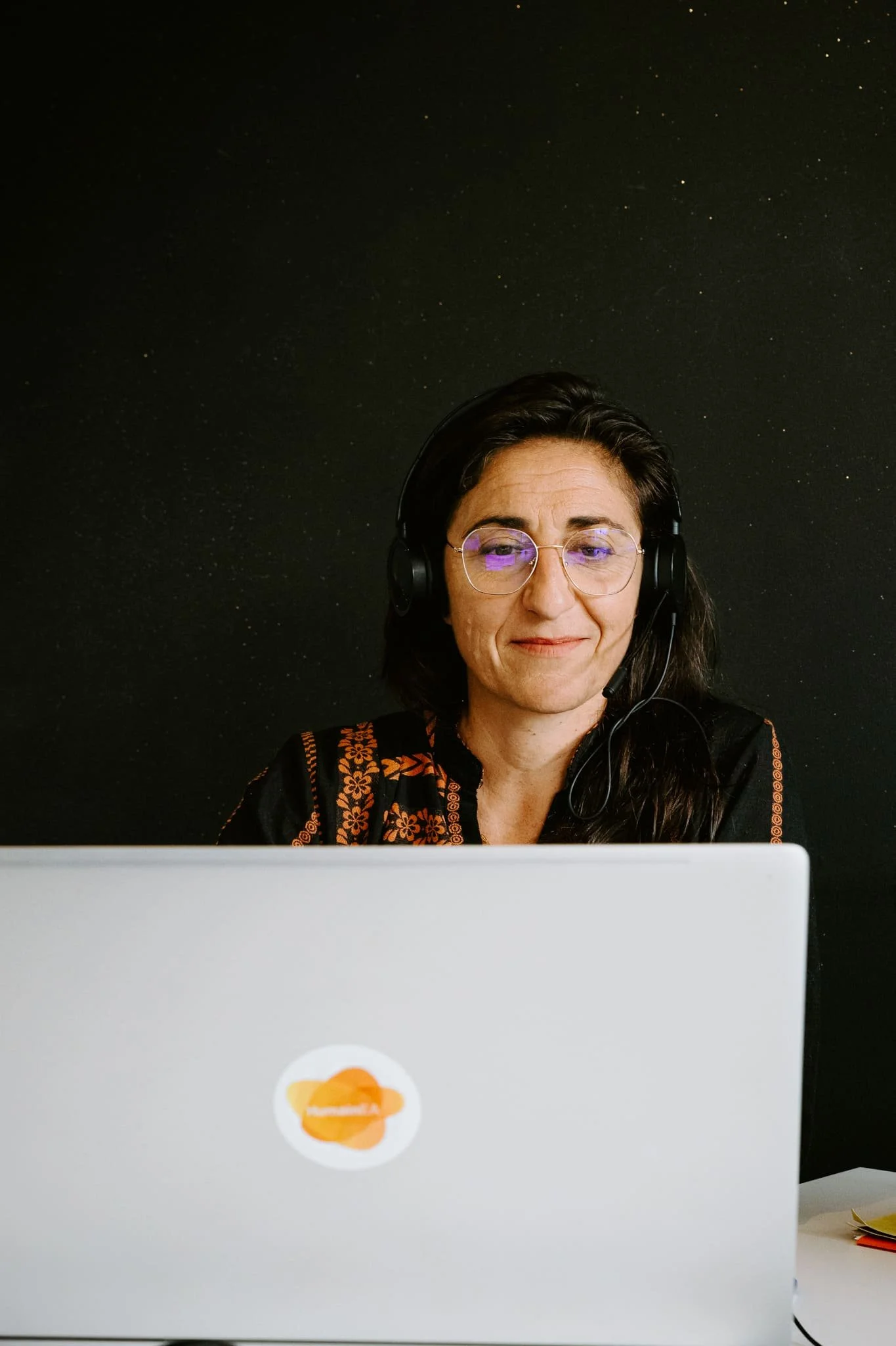 Photo prise dans les bureaux de une entreprise RH d'insertion marseillaise. On y voit une femme souriante portant un casque avec micro, portant des lunettes, assise derrière un ordinateur portable blanc, devant un mur sombre.