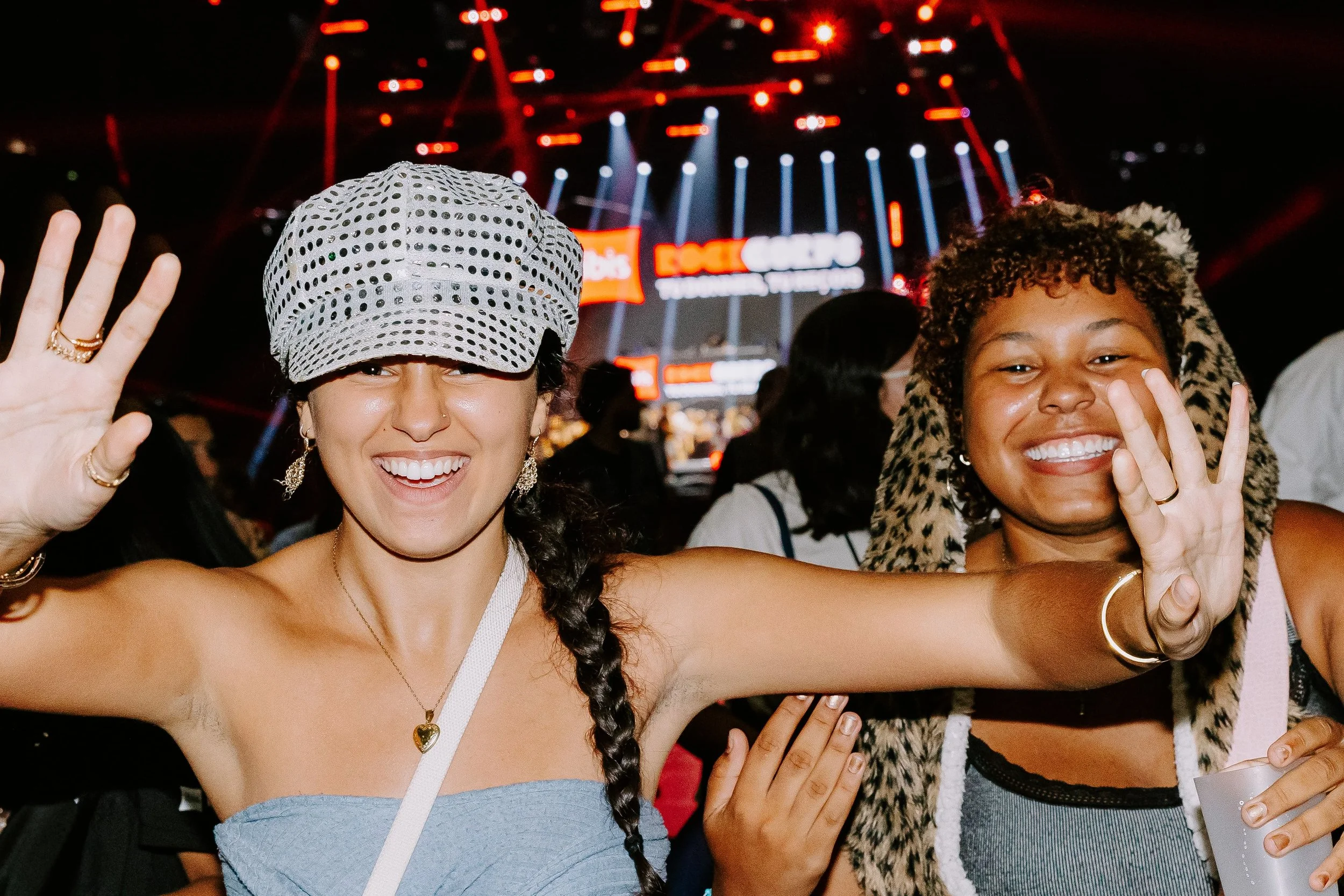 Deux femmes souriantes à un événement, faisant un signe de paix avec leurs mains, en arrière-plan un écran lumineux avec des textes, lumières et scène de concert.