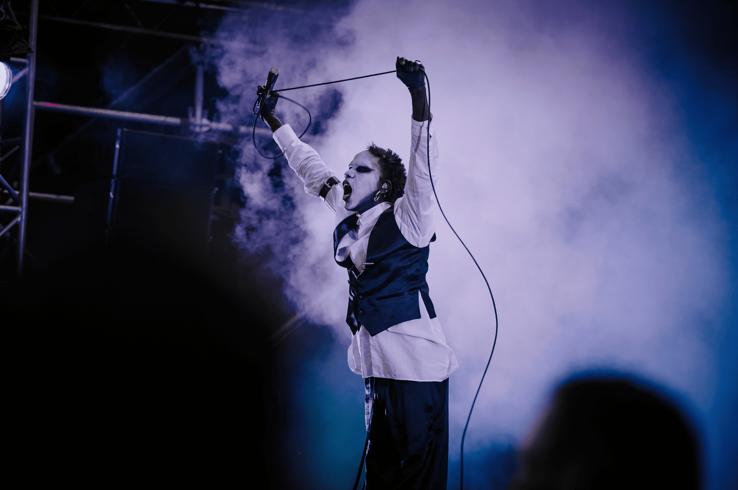 Un chanteur en costume noir et blanc avec du maquillage sombre, tenant un micro et une rallonge de micro, se produisant sur scène avec des effets de fumée sur fond sombre.
