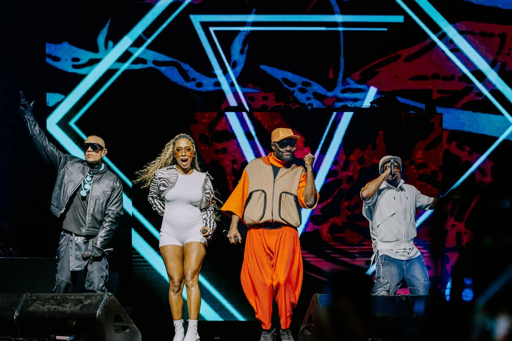 Quatre artistes ou performeurs sur scène avec un fond lumineux coloré en forme de géométrie abstraite, vêtus de tenues modernes et expressives, en pleine performance ou émission musicale.