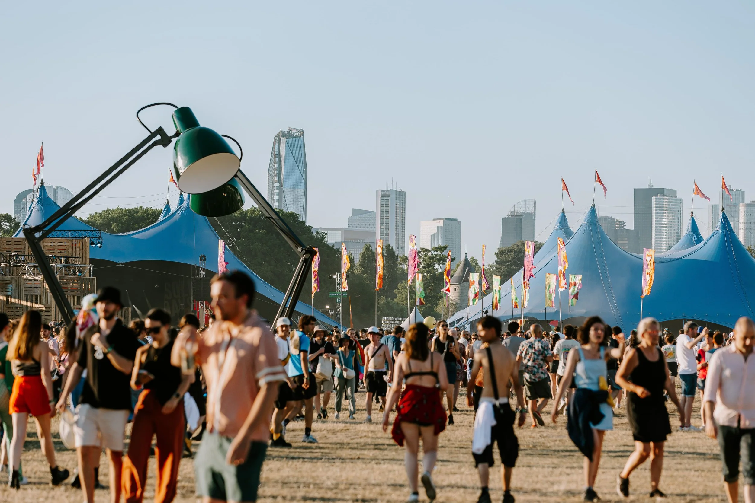 Festival en plein air avec des stands et une foule de personnes, des tentes bleues, drapeaux colorés, et un skyline de gratte-ciel en fond.