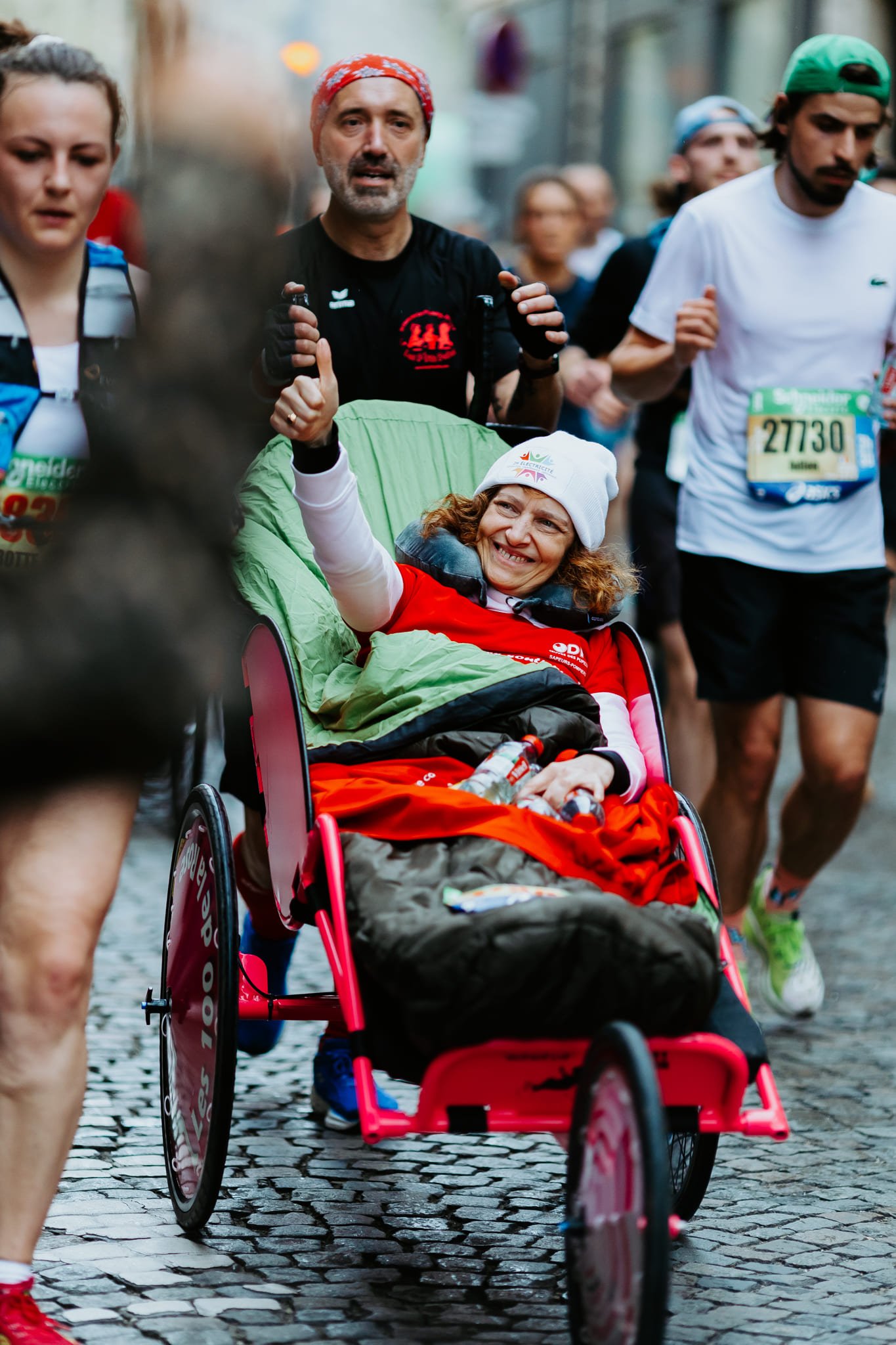 Femme en fauteuil roulant souriante, entourée de coureurs, durant le marathon de Paris. La femme porte un bonnet blanc et une veste rouge, elle fait un geste de victoire avec la main.