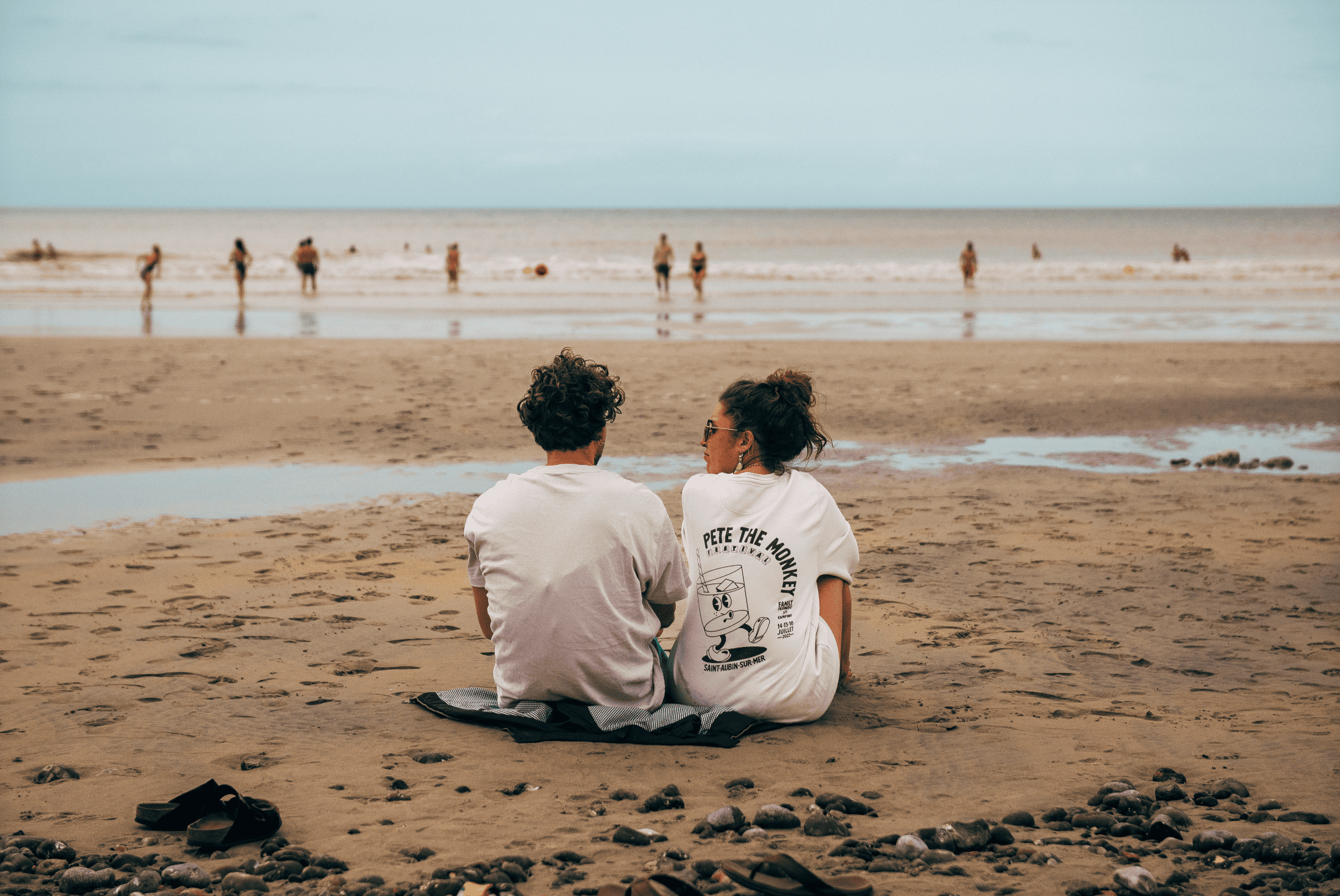 Deux personnes assises sur la plage face à l'océan, regardant l'horizon, avec d'autres personnes au loin dans l'eau, ciel nuageux, sable et galets au premier plan.