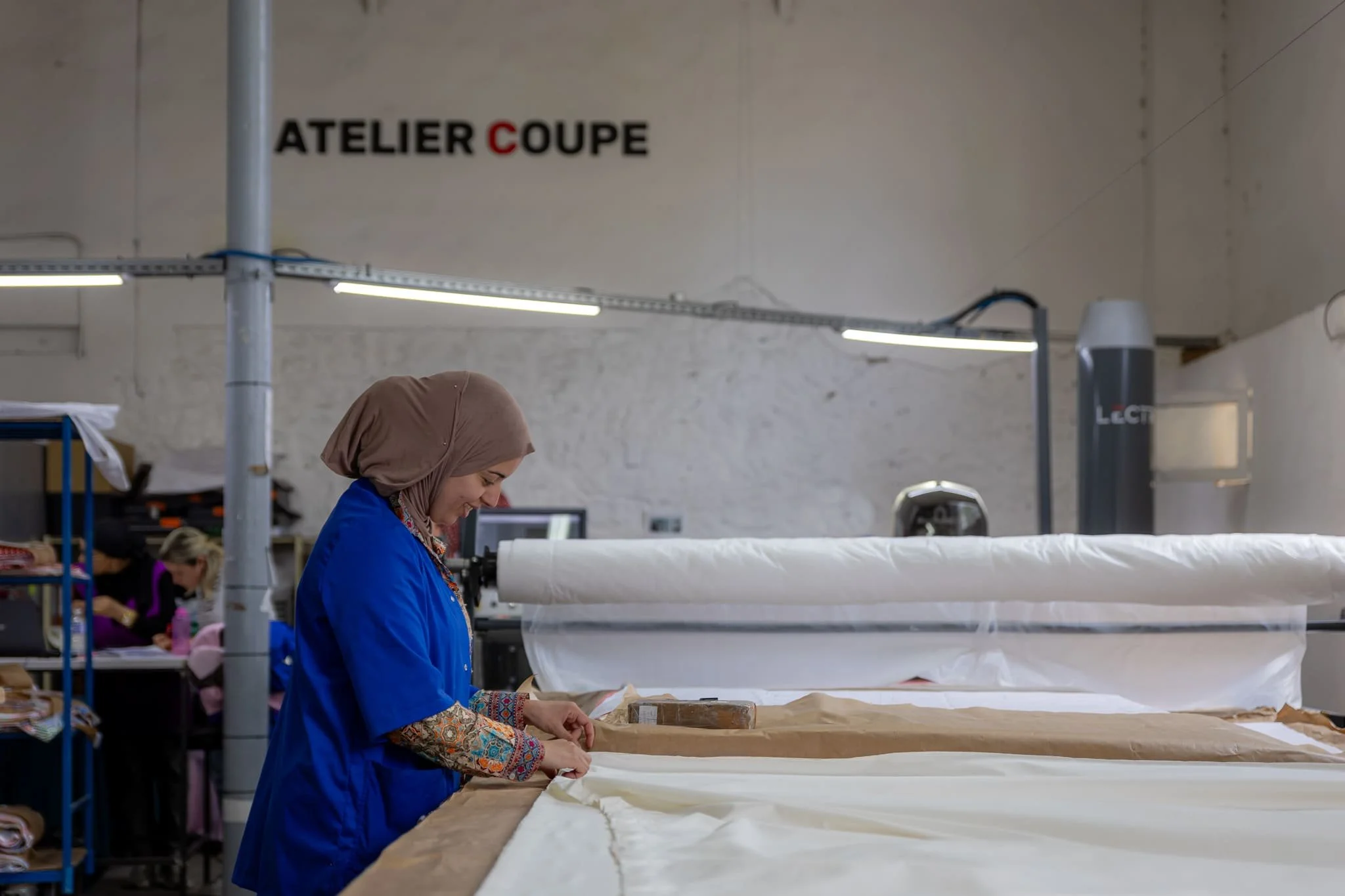 Reportage photo dans un atelier textile marseillais. On y voit une femme portant un hijab beige couper du tissu sur une grande table. Sur le mur en arrière plan, est écrit 'ATELIER COUPE'.