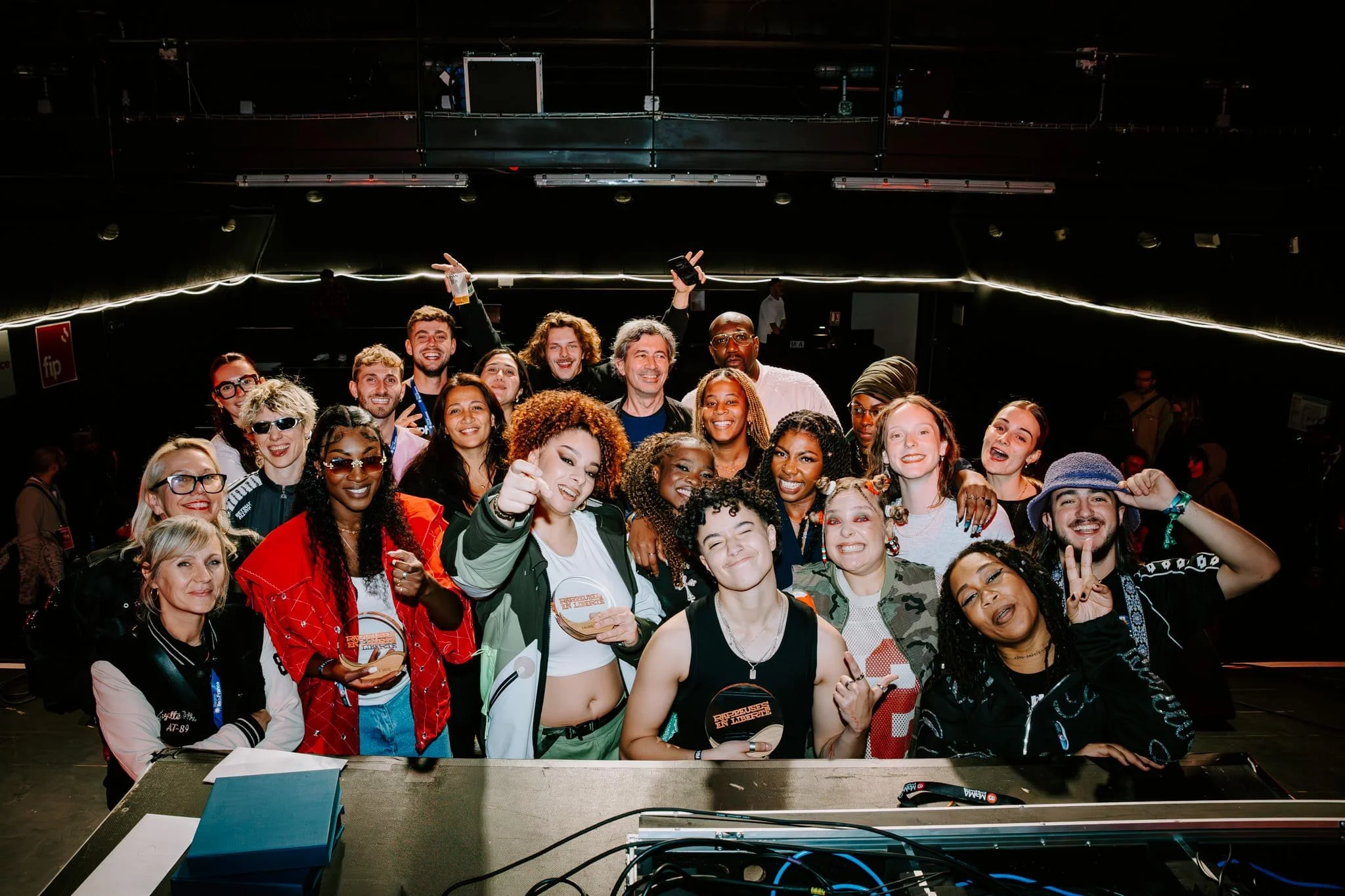Photo d'un groupe de personnes souriantes posant sur scène pour une photo dans une salle sombre, avec du matériel technique au premier plan. Prise à l'occasion de la finale du dispositif Rappeuses en Liberté.