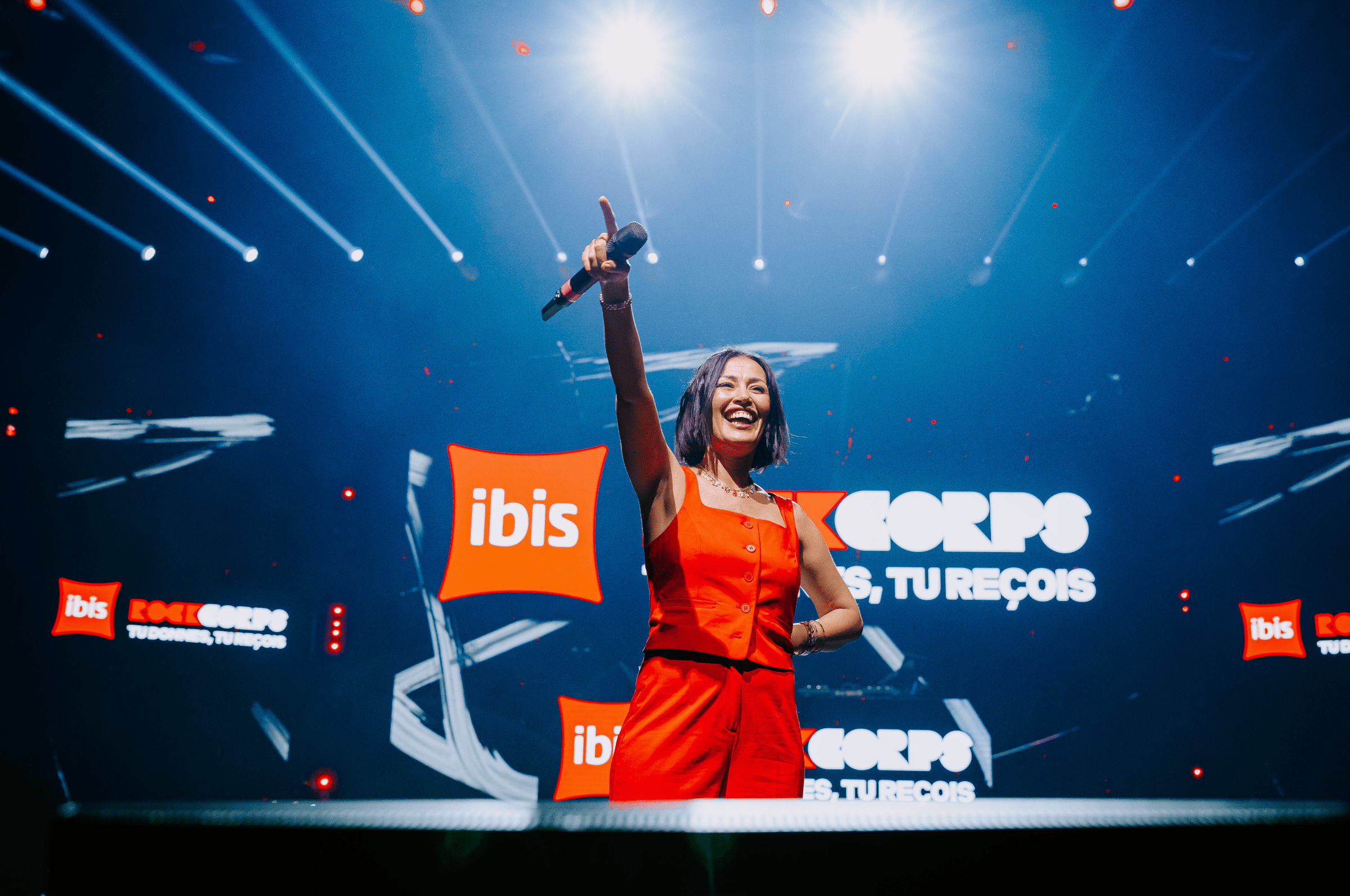 Photo de l'animatrice TV et radio Karima Charni, souriante, brandissant un micro face à la foule. En arrière plan des écrans affichant les logos de l'événement : Ibis Rockcorps.