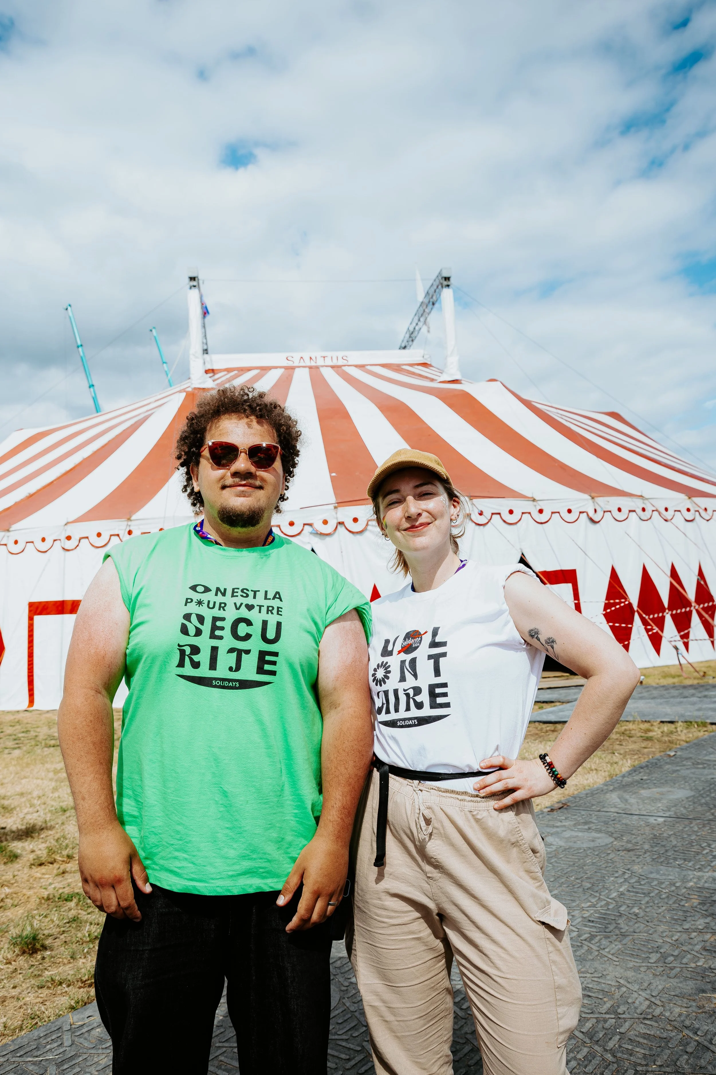 Deux personnes posent devant une grande tente de cirque avec un ciel nuageux, un homme avec des cheveux bouclés, lunettes de soleil vertes, t-shirt vert, et une femme avec un chapeau beige, t-shirt blanc et pantalon beige, dans un environnement en plein air.