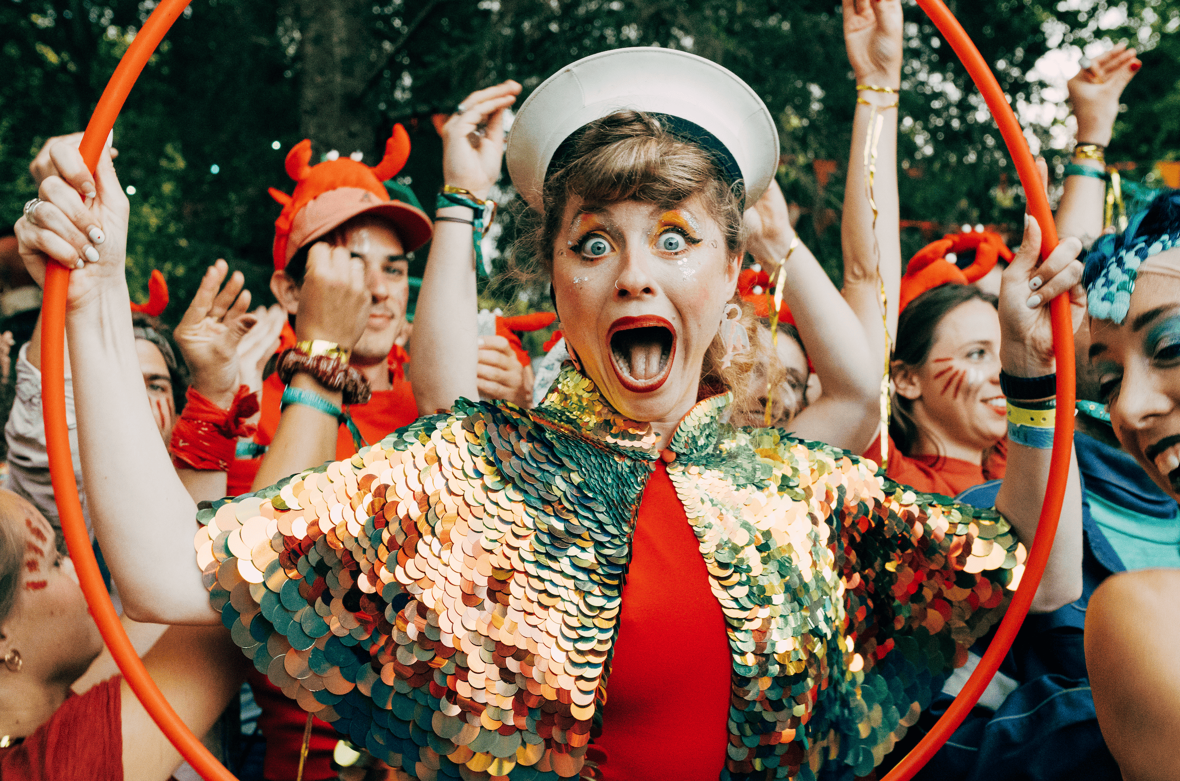 Une femme habillée de sequins colorés, avec un chapeau blanc, criant d'excitation lors d'une fête costumée, entourée de personnes avec des costumes colorés et des maquillages festifs.