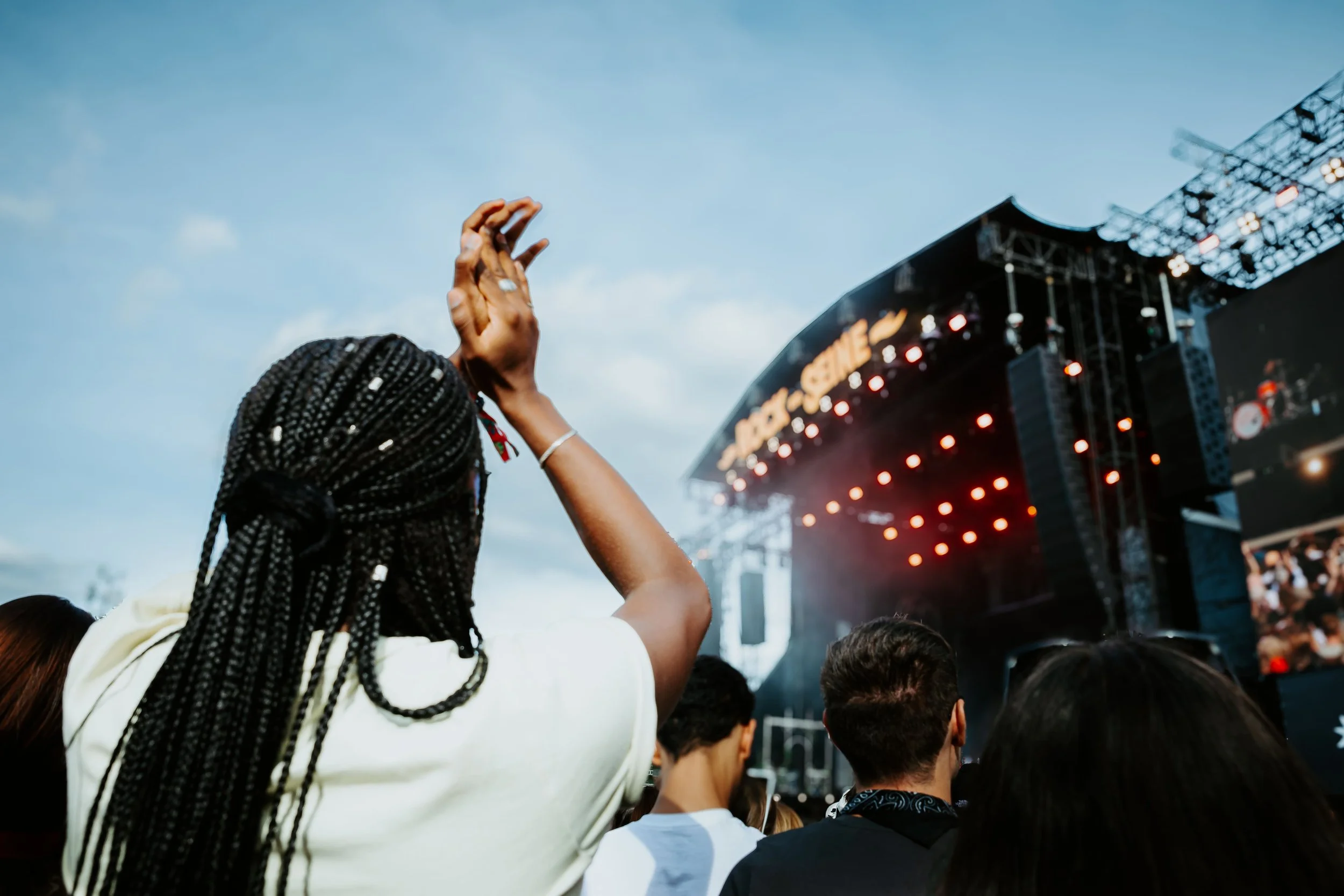 Un public assiste à un concert en plein air, avec une scène éclairée par des lumières rouges et un ciel en fin d'après-midi ou début de soirée.