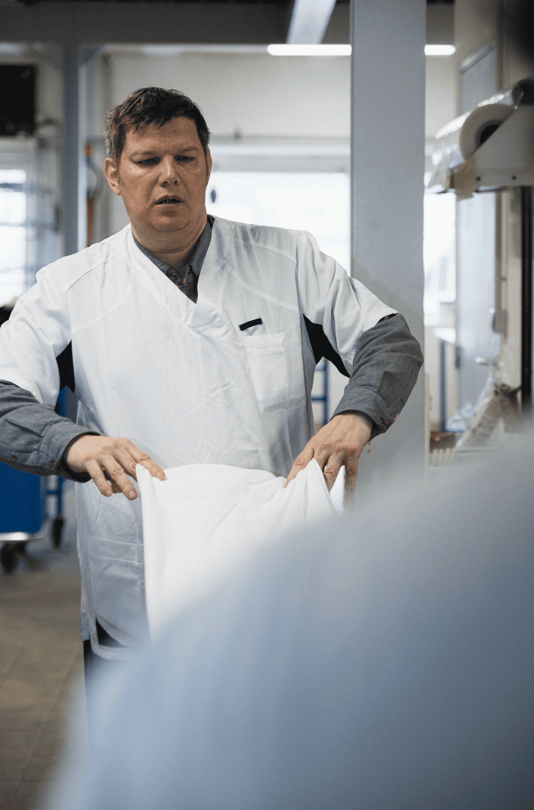 Photo prise au seine de l'ESAT les Glycines à Marseille. Un homme en uniforme de travail, travaillant dans un atelier de blanchisserie, plie un drap avec son collègue qu'on aperçoit de dos au premier plan.