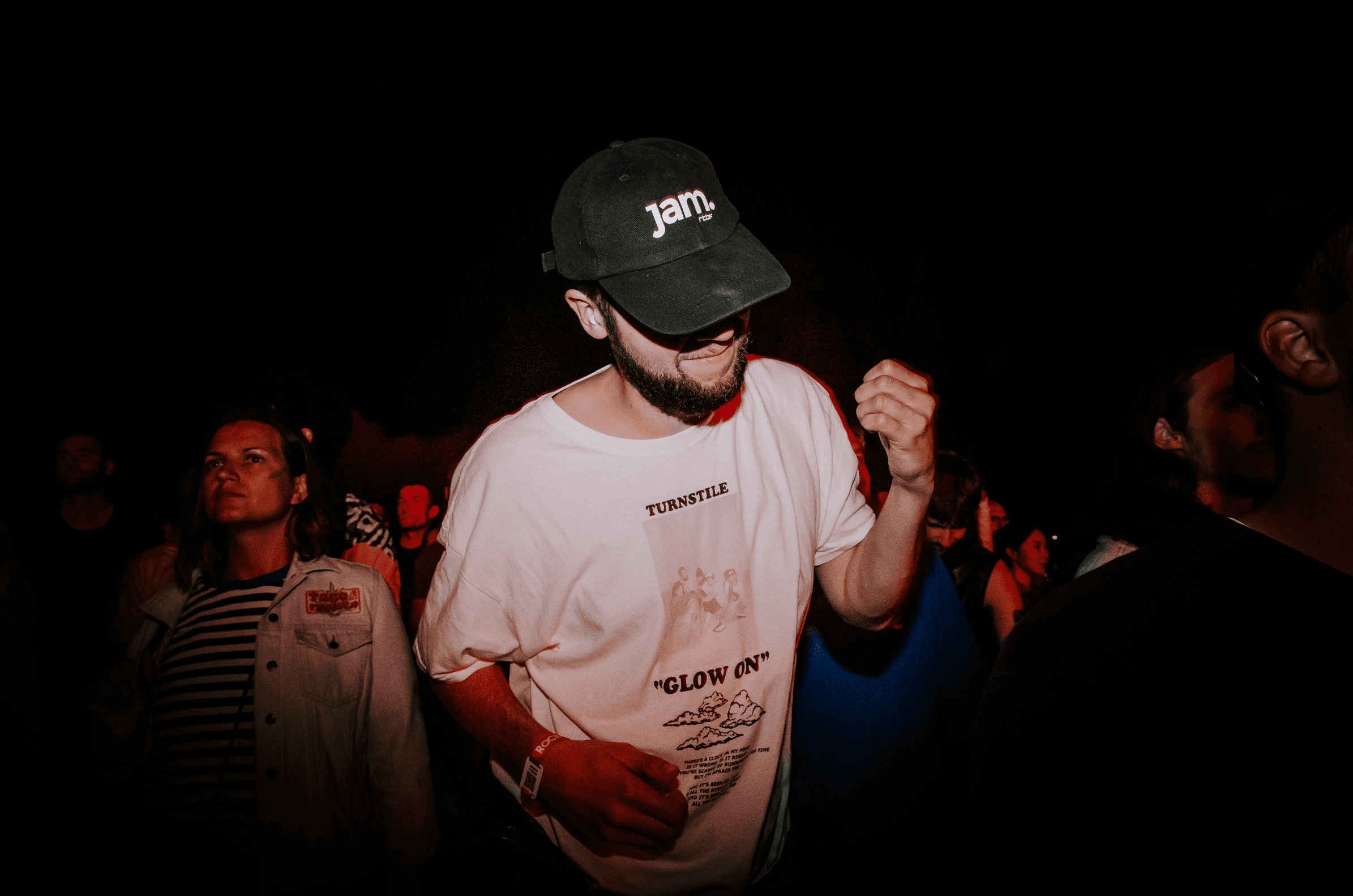 Un homme portant une casquette noire, lunettes et un t-shirt blanc avec un graphique, semble danser ou bouger au rythme de la musique, dans un environnement sombre avec d'autres personnes autour.