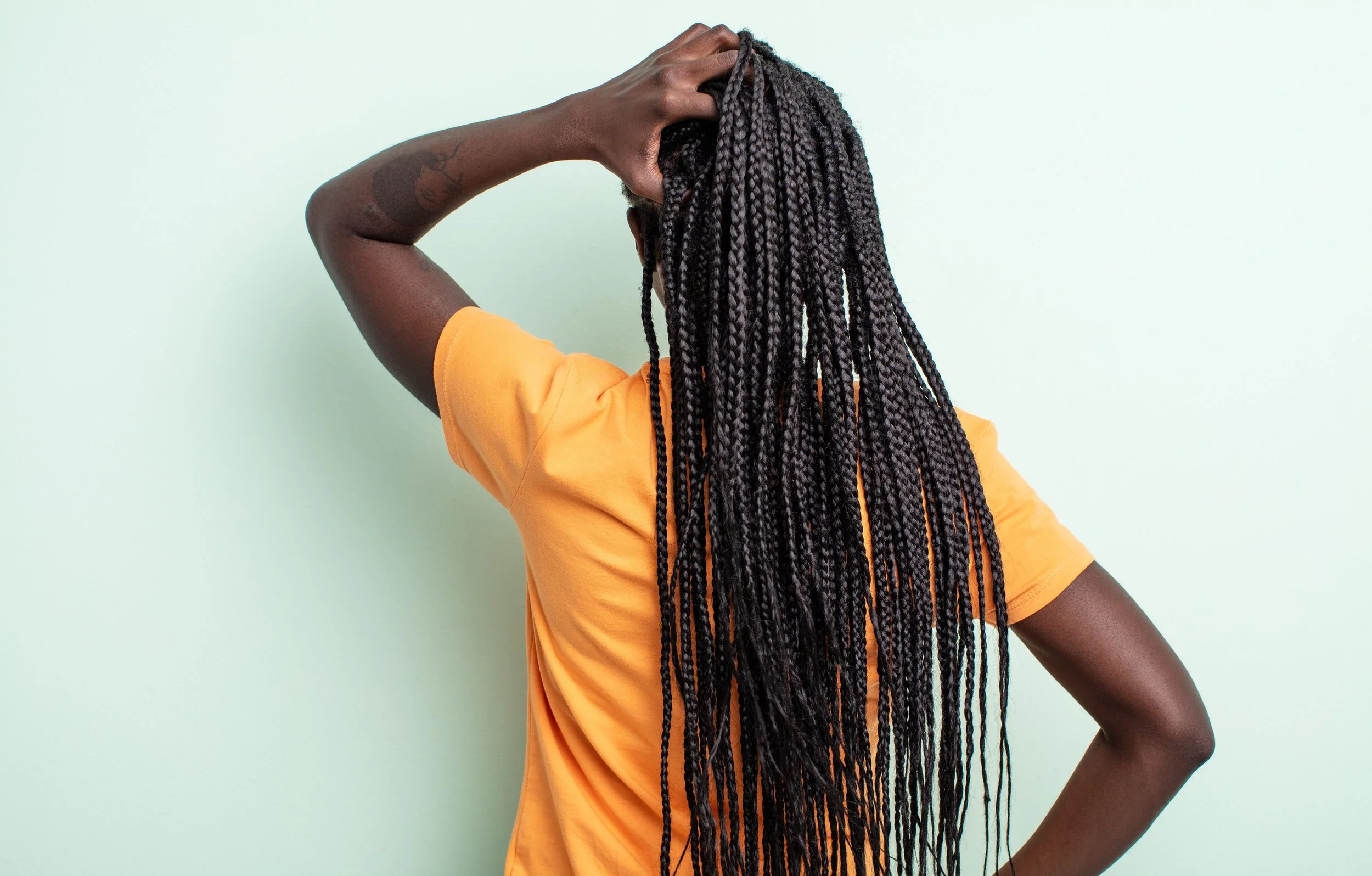 Femme avec coiffure en longues tresses de détail, portant un t-shirt orange, posant avec une main sur la tête, fond vert clair.