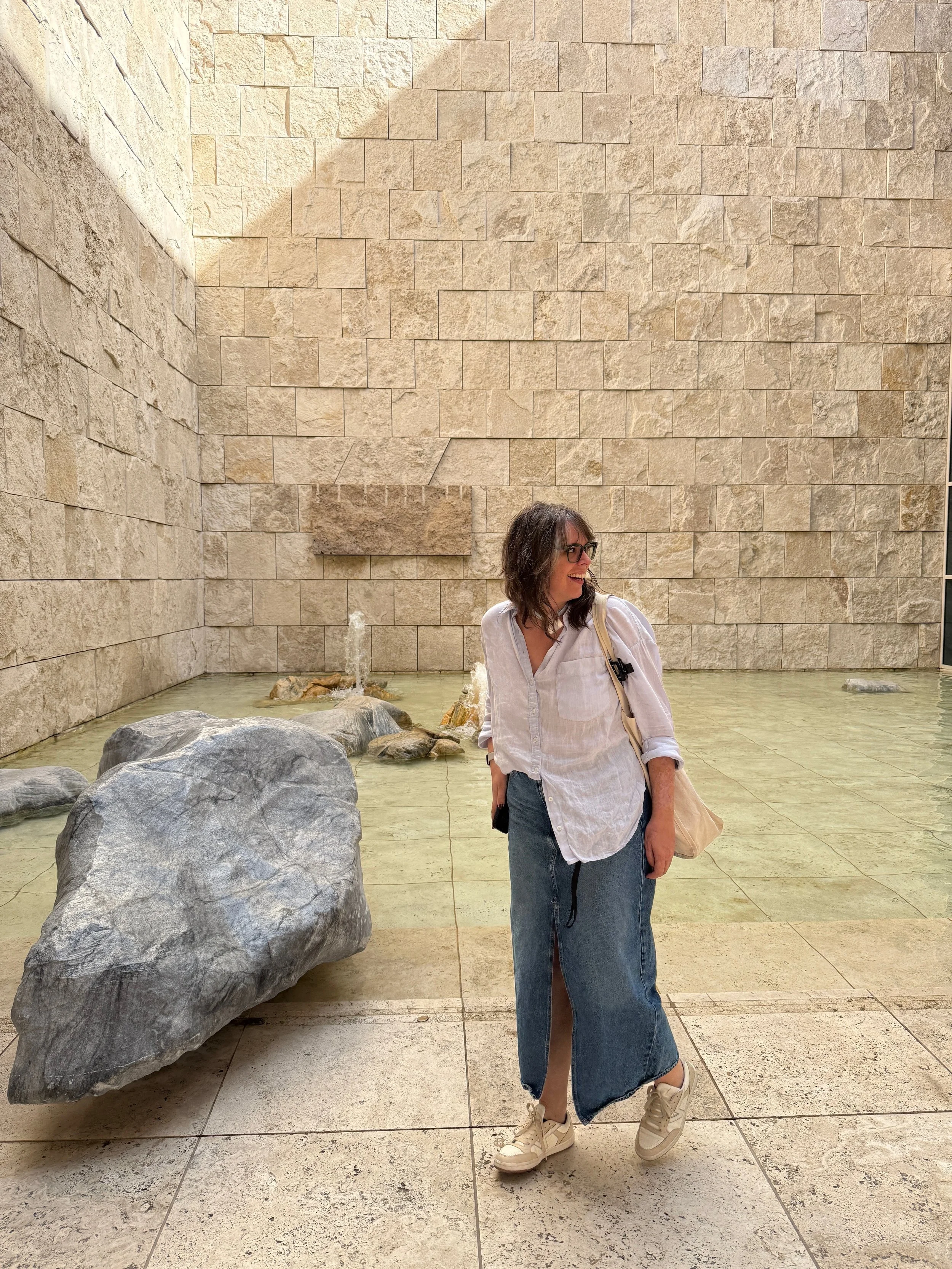 A woman wearing glasses, a white shirt, and a long denim skirt standing in front of a stone wall with water and rocks, smiling and looking to her left.