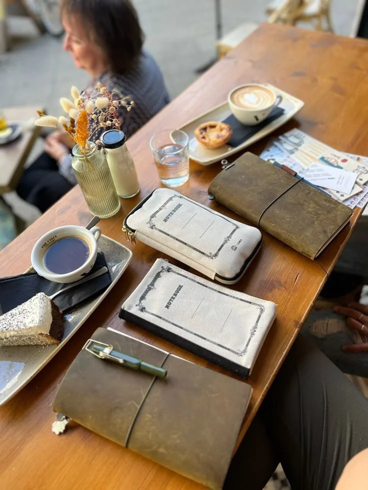 A wooden table with assorted items including two notebooks, a leather-bound journal, a pencil case, a cup of coffee, a slice of cake, a glass of water, a cup of latte, a small vase with dried flowers, and a woman sitting in the background.