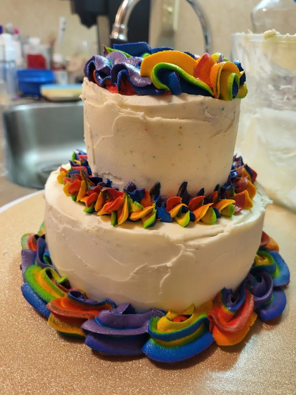 A two-tiered cake decorated with multicolored icing in rainbow swirl patterns around the base and top edges, featuring a smooth white frosting surface.