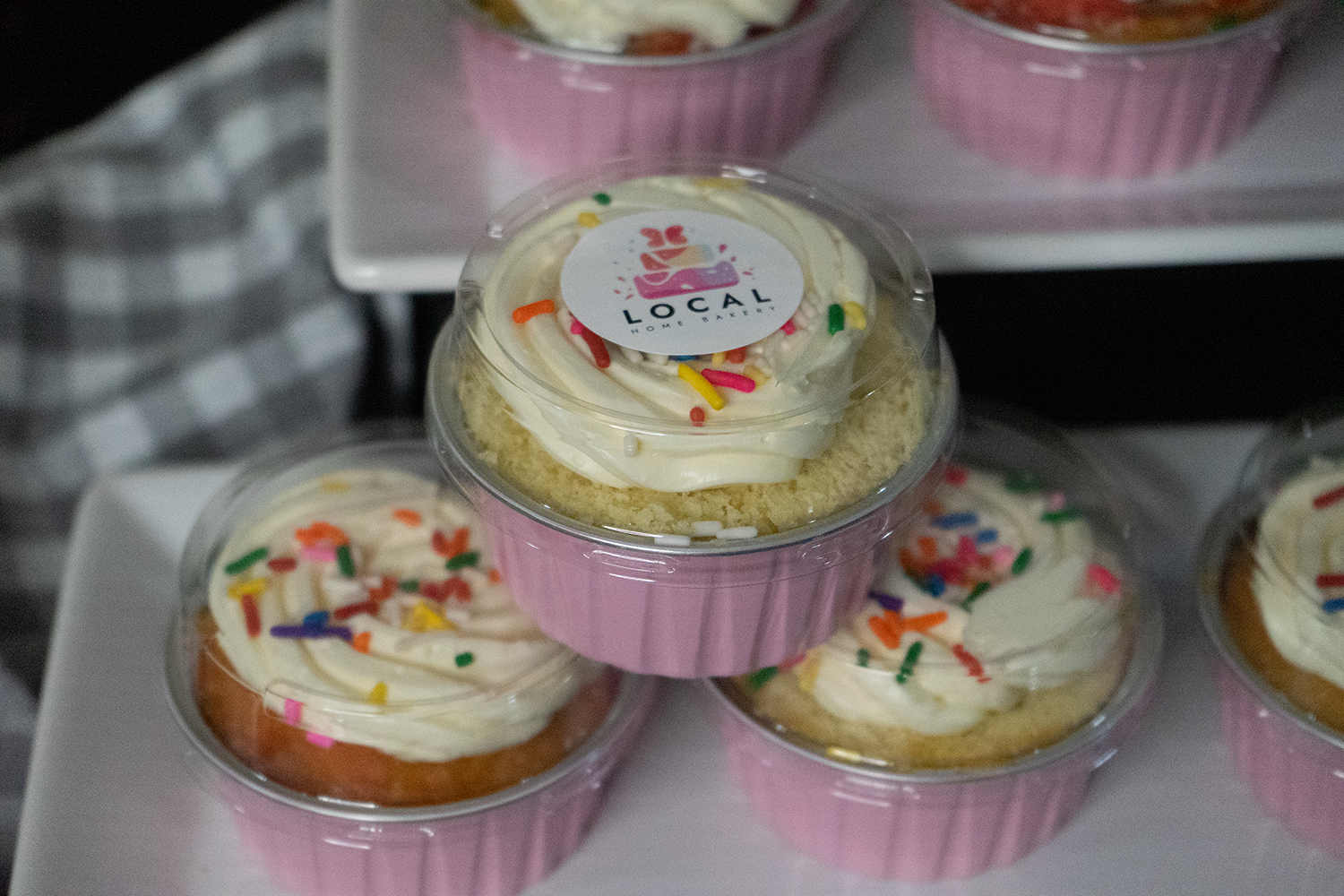 Mini cakes with white frosting and colorful sprinkles in pink paper liners, arranged on a white tray.